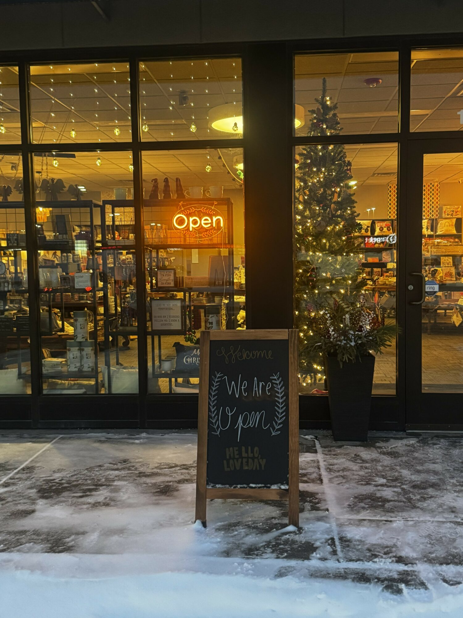 Store window with Christmas tree and an 'Open' sign, snow on ground, and a chalkboard sign outside.
