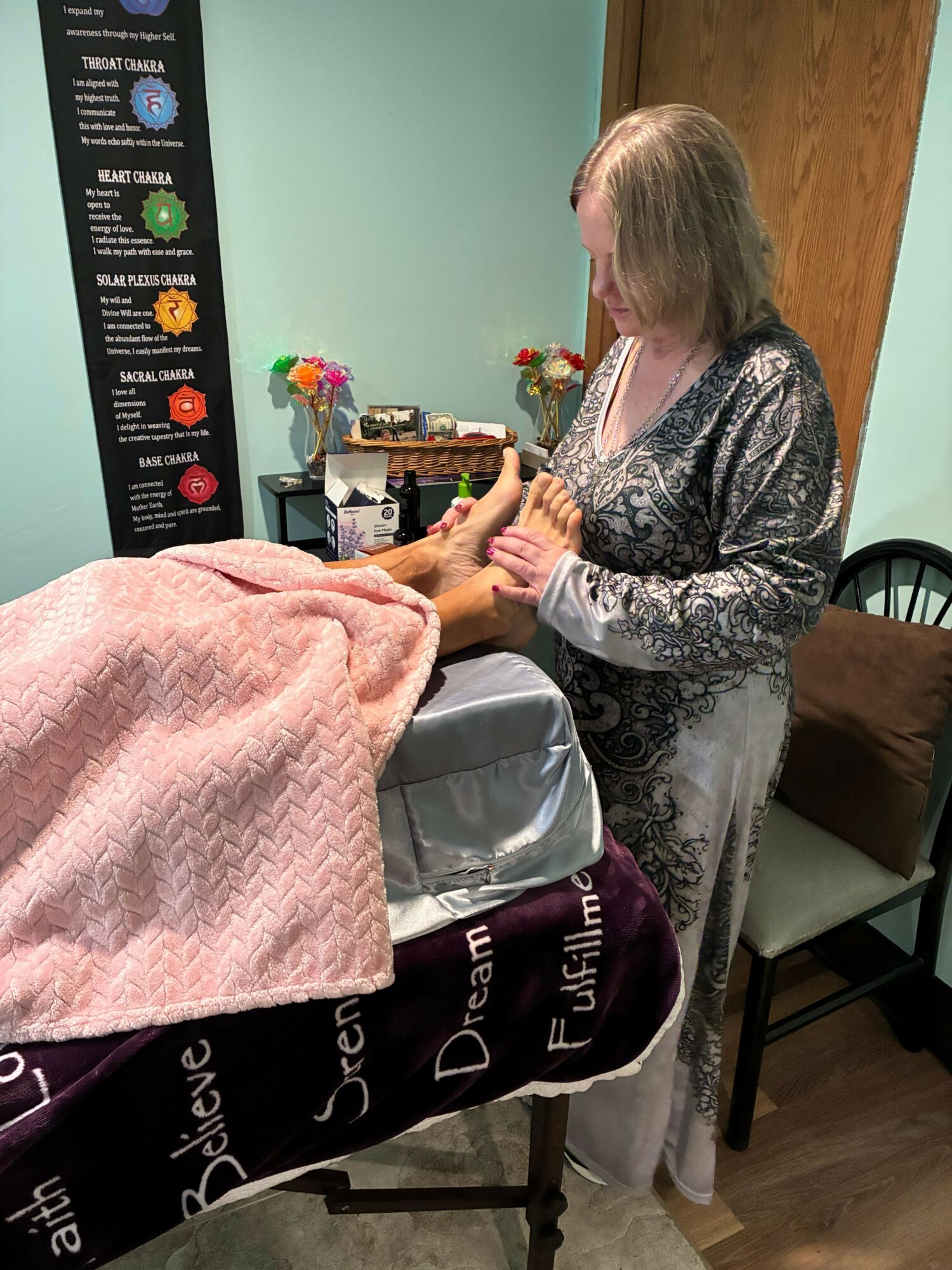 Woman with long hair in patterned dress hands a gift to a person seated at a table, pink blanket draped over the table.