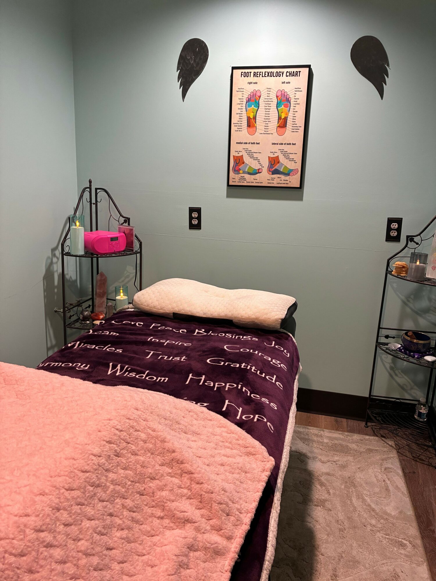 A bed with a pink blanket and purple bedspread in a room with a framed colorful artwork on the wall, a small table, and a pink object.