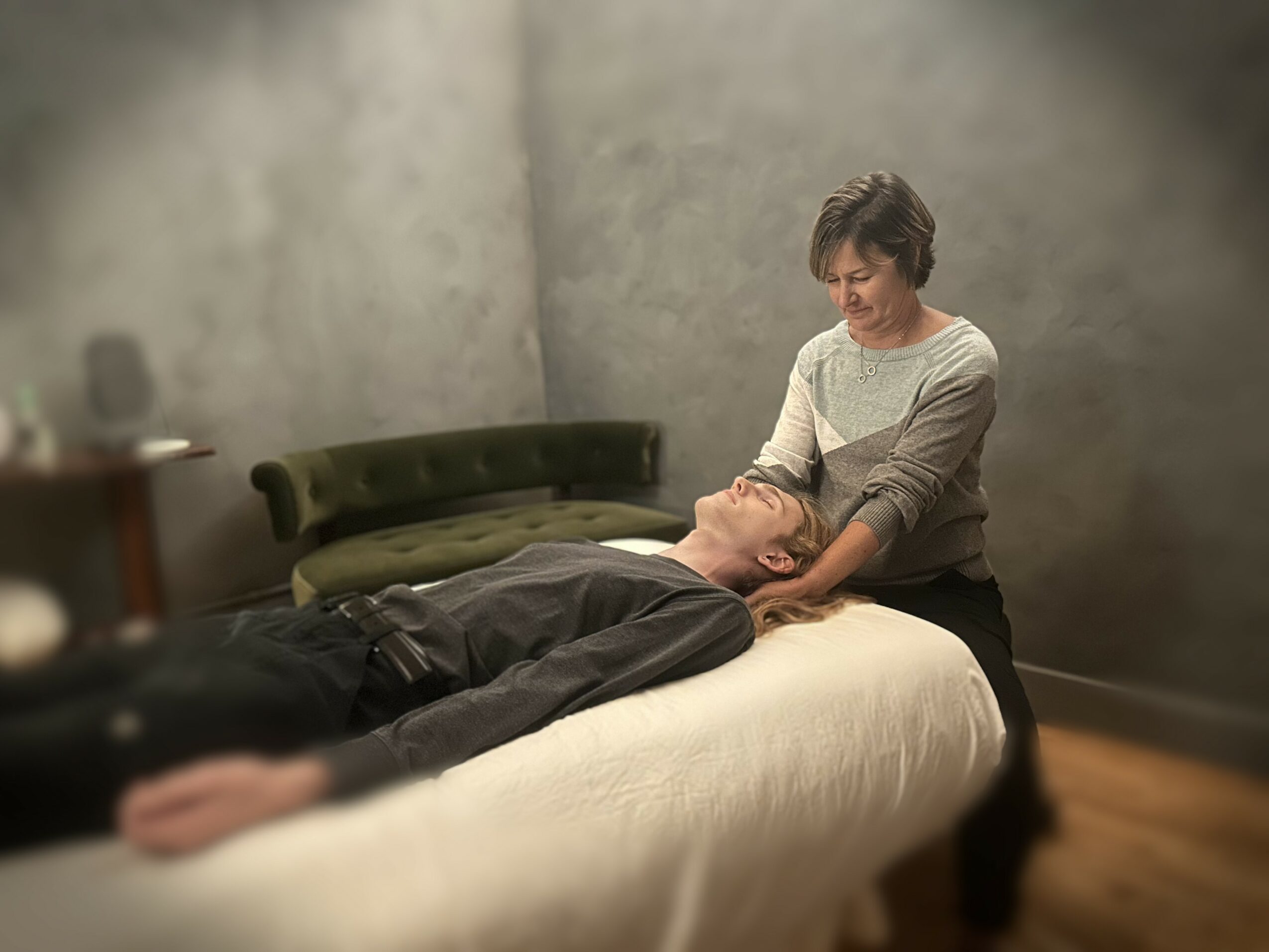 Woman providing a massage to a man lying on a bed in a room with gray walls and a green sofa.