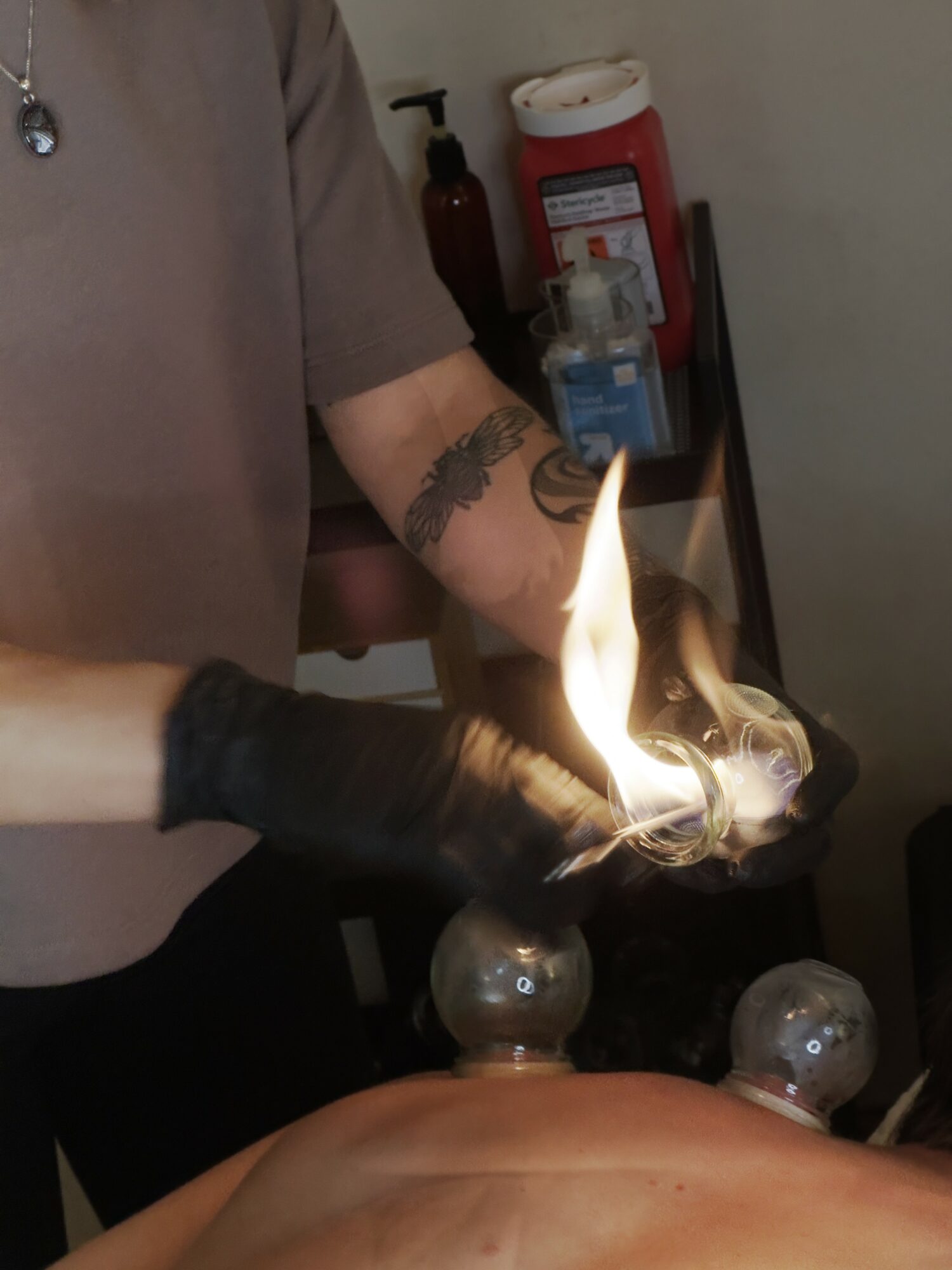 Person wearing gloves holding a flame over a glass object, with a tattooed arm visible, in a room with safety equipment.