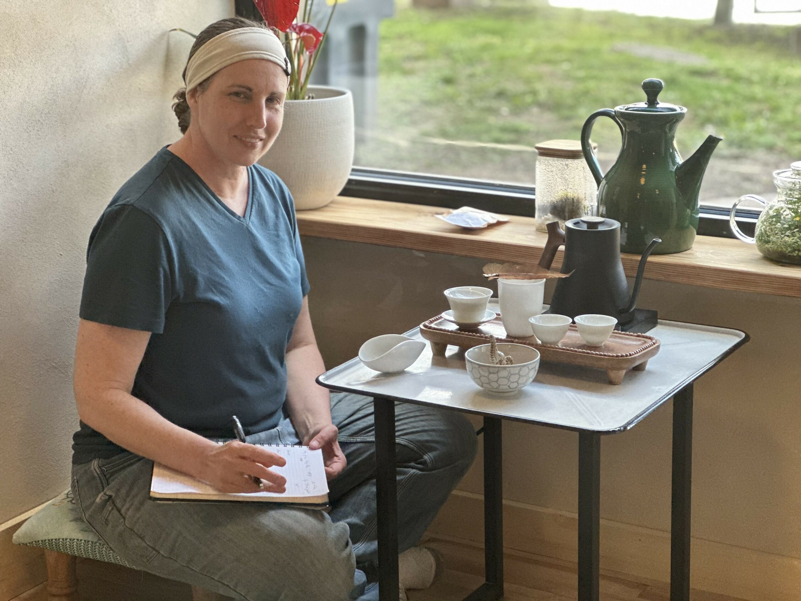 Woman sitting by window with tea set and plants, writing in notebook, natural light, indoor setting.
