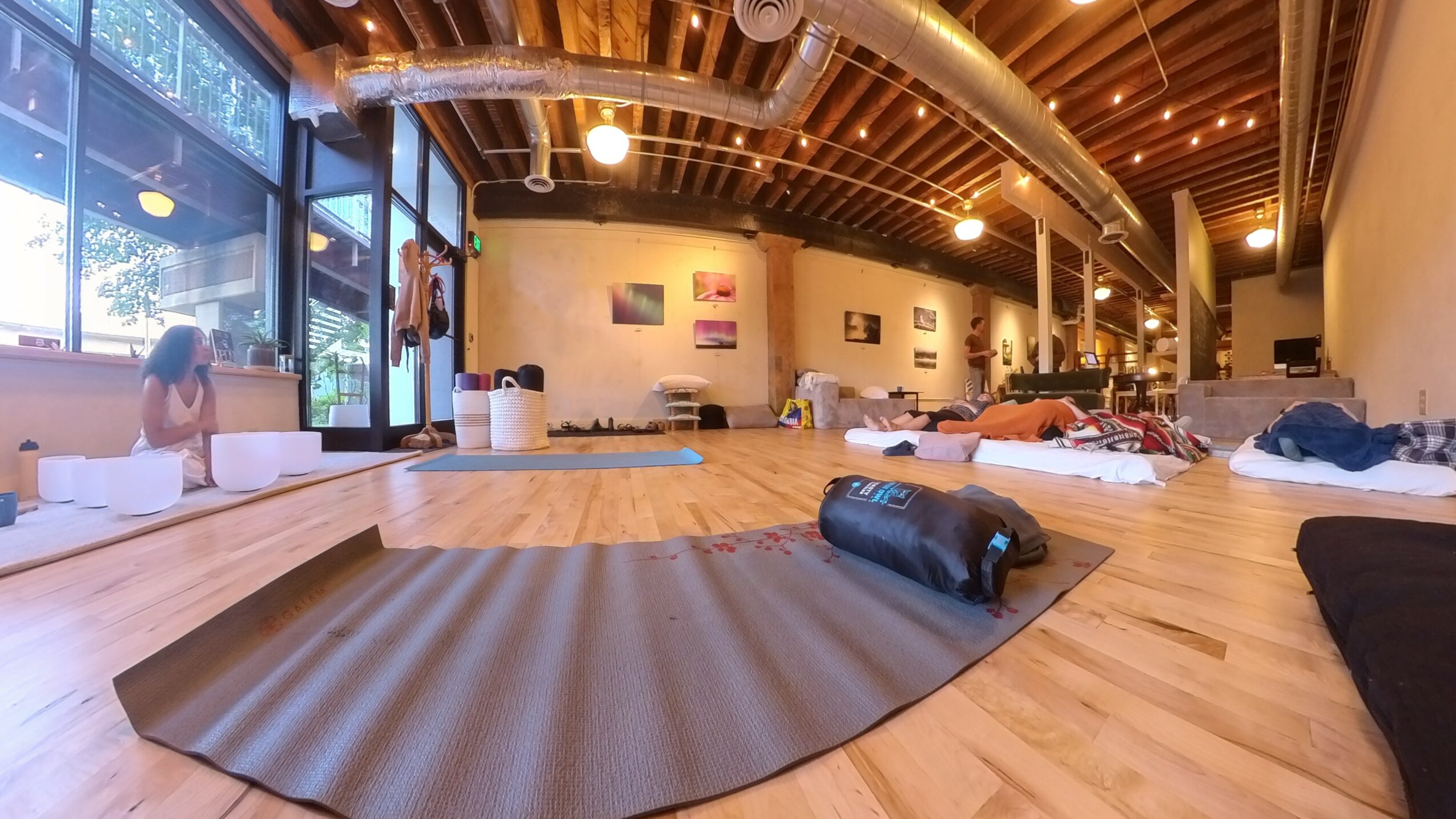 Yoga mats and blankets arranged on a wooden floor in a spacious, well-lit studio with large windows and artwork on walls.