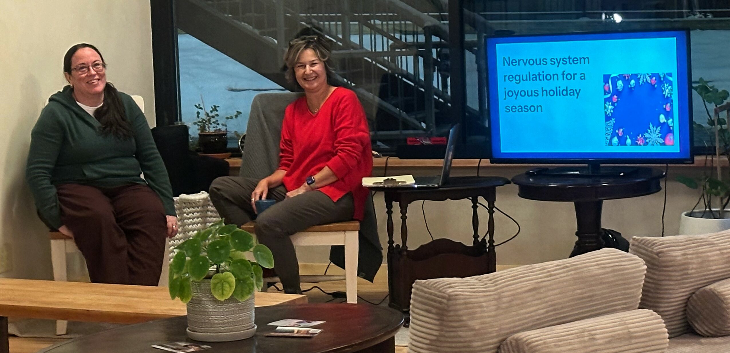 Two women sitting on chairs in a room with a screen displaying a presentation slide, and a table with a plant in the foreground.