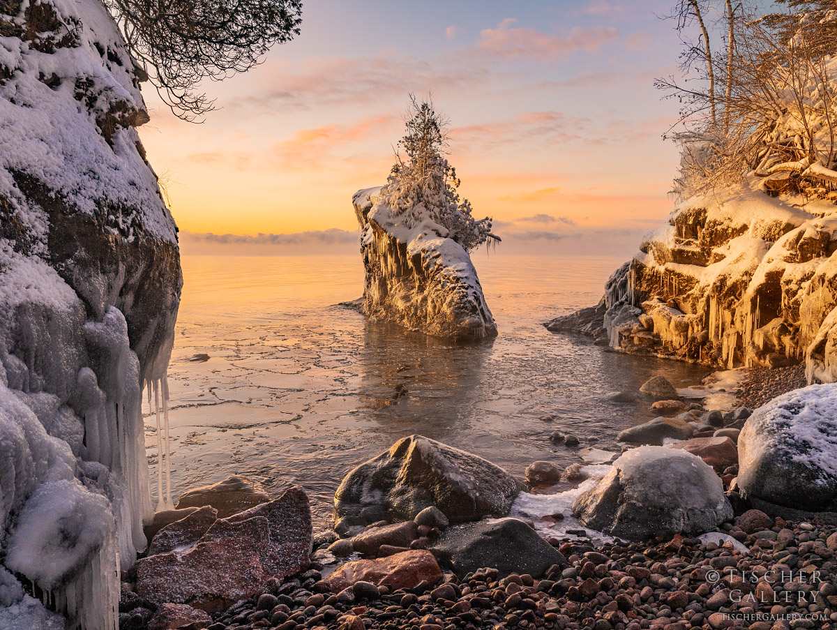 Snow-covered rocks and trees by a frozen shoreline at sunset with a large ice formation in the water.