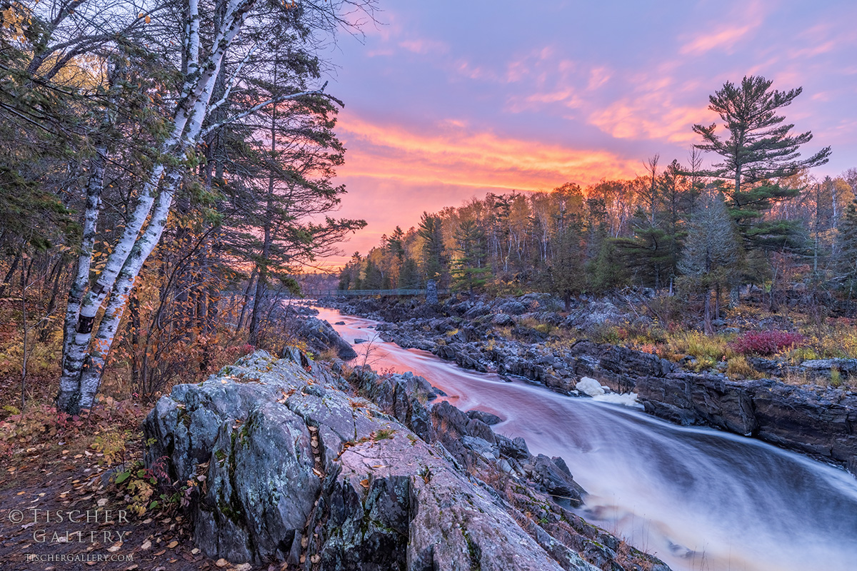 River flowing over rocks with trees and colorful sunset sky in background.