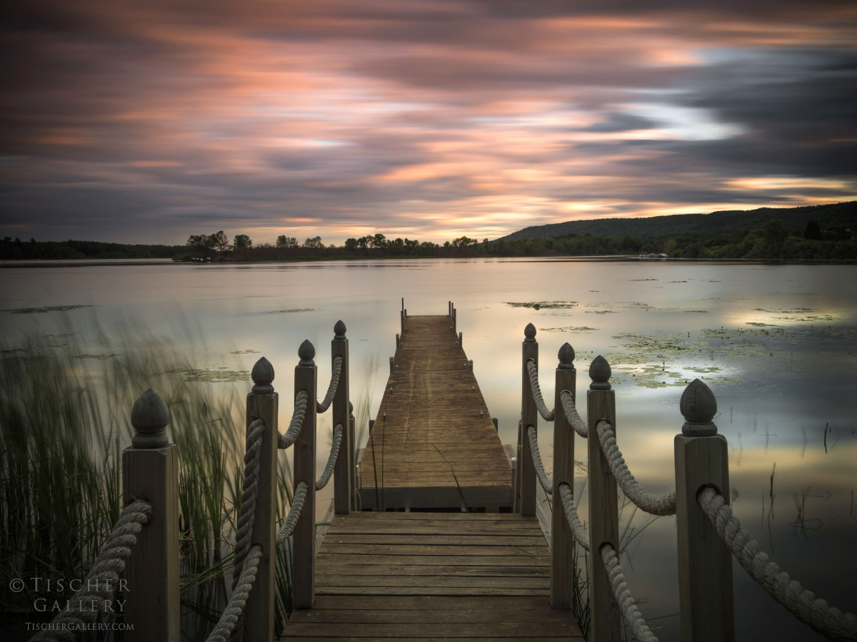 Wooden dock extends into a calm lake with a cloudy sky at sunset, trees in the distance, and water reflections.