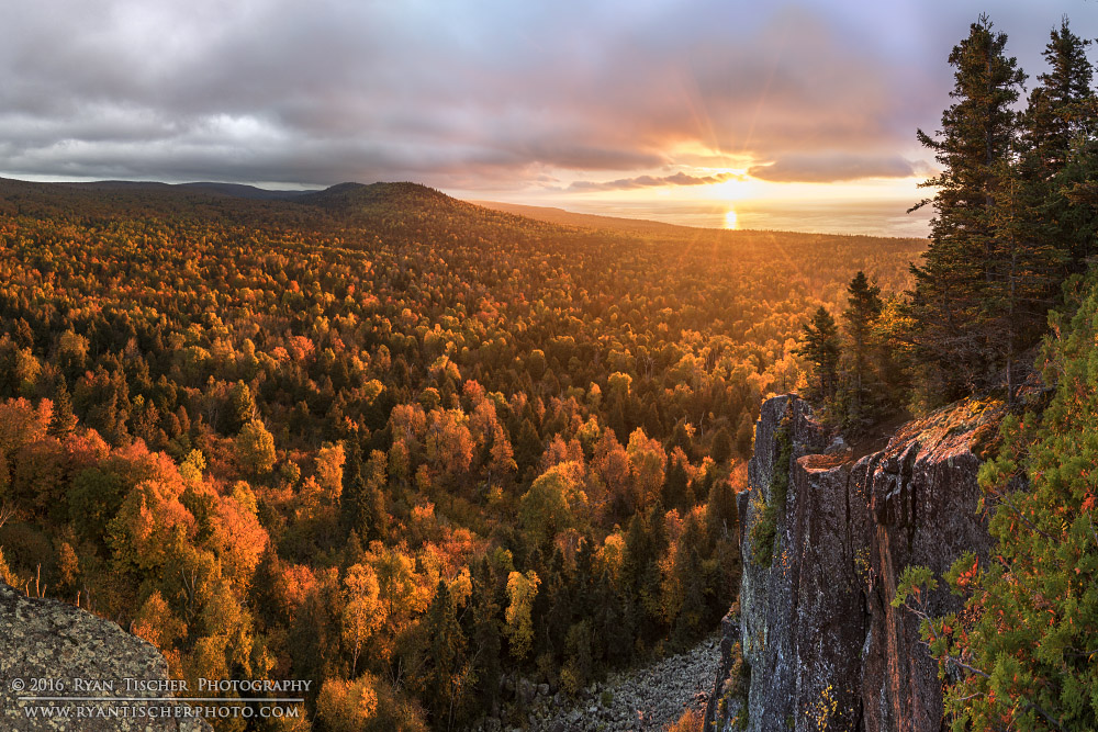Sunset over a forested landscape with a waterfall on the right, orange and green trees, cloudy sky, and distant hills.