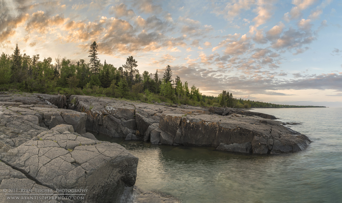 Rocky shoreline with trees and a cloudy sky at sunset, calm water in the foreground.