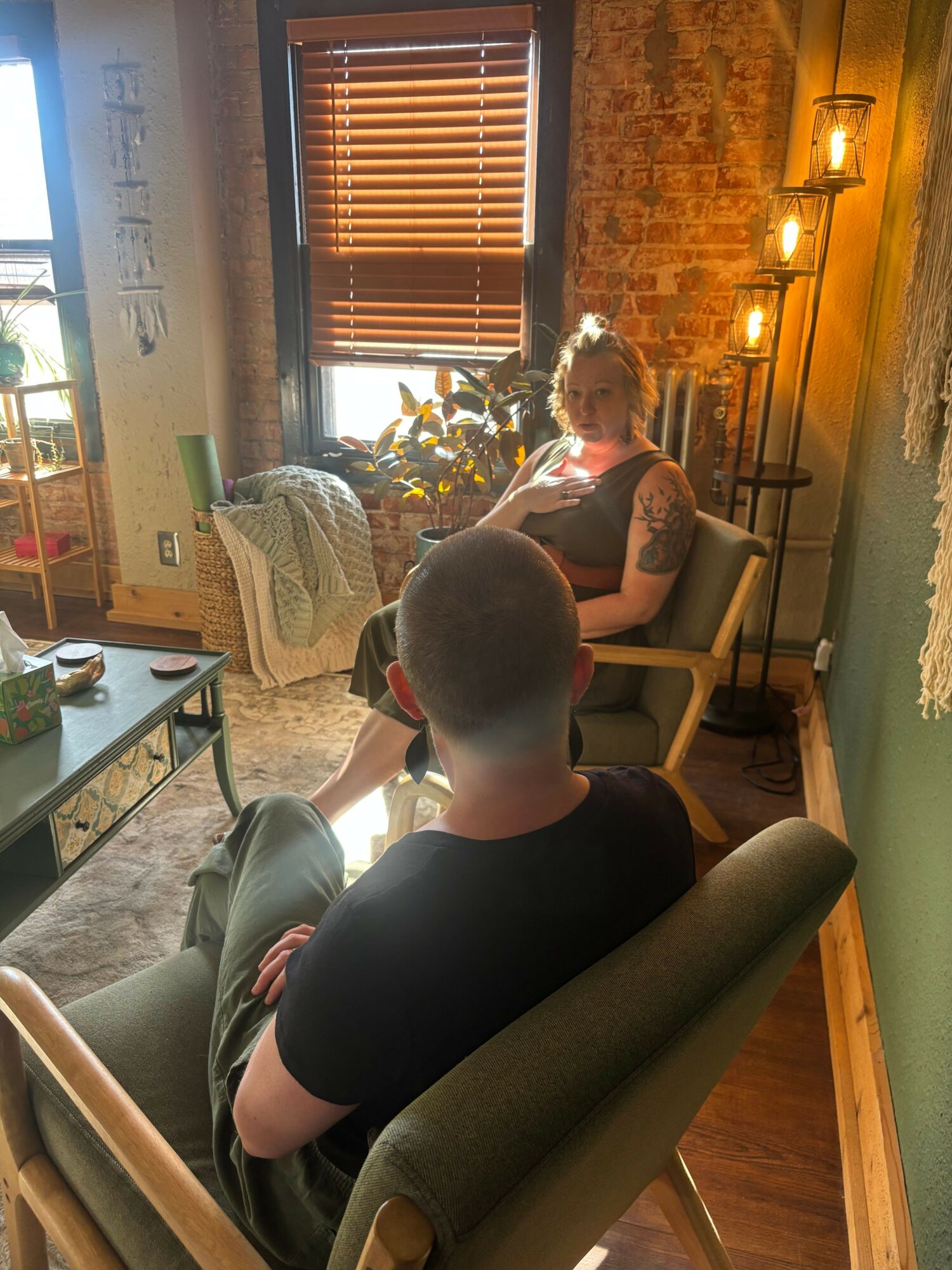 People sitting and standing in a room with wooden walls and window blinds, engaging in conversation or activity.
