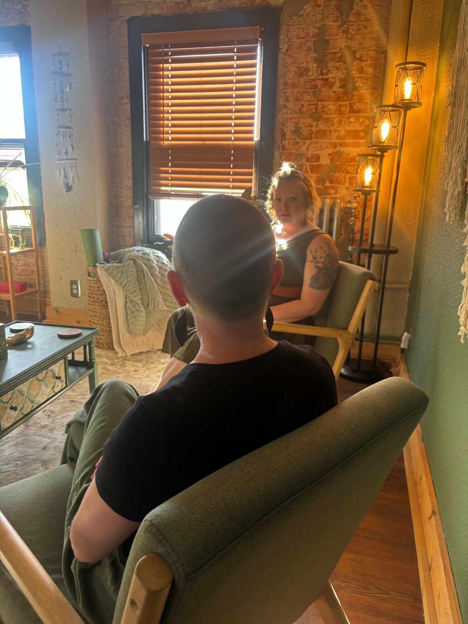 Person with shaved head sitting on a chair in a cozy room with brick wall and window blinds.