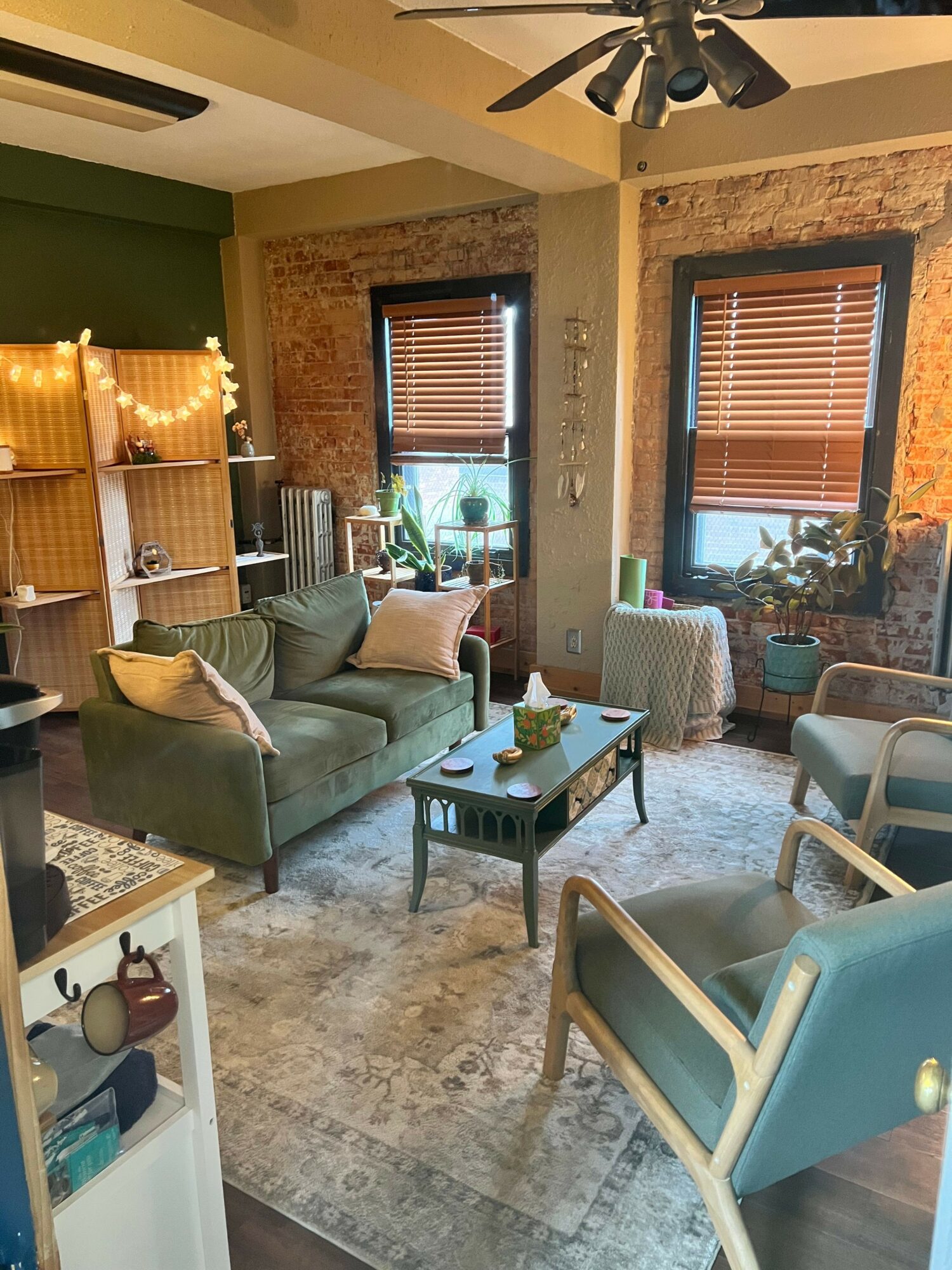 Living room with green walls, brick wall, sofa, armchair, coffee table, and large windows with blinds.