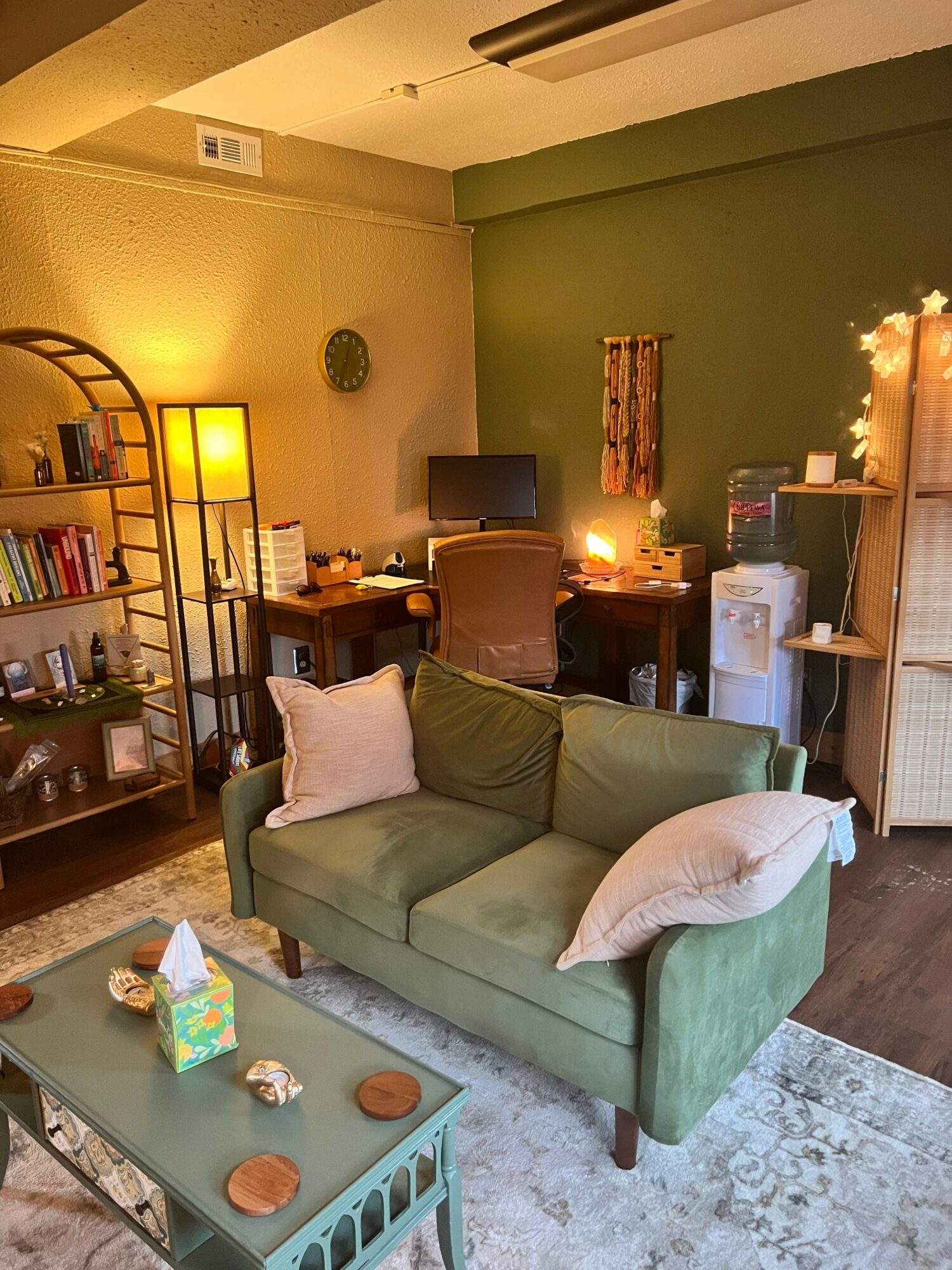 Living room with green wall, sofa with pillows, bookshelf, and a coffee table with items, viewed from the side.