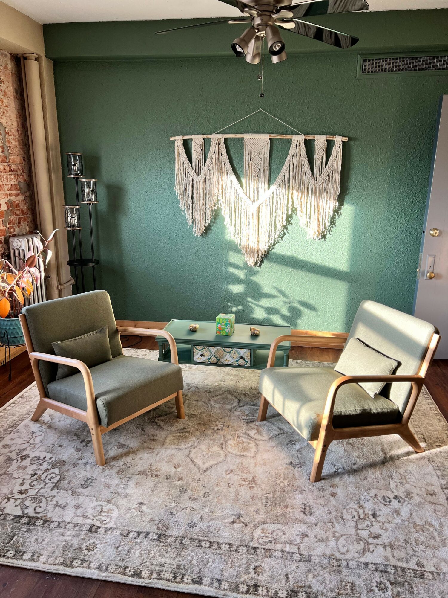 Living room with green wall, hanging wall art, two chairs, a small table, and a patterned rug.