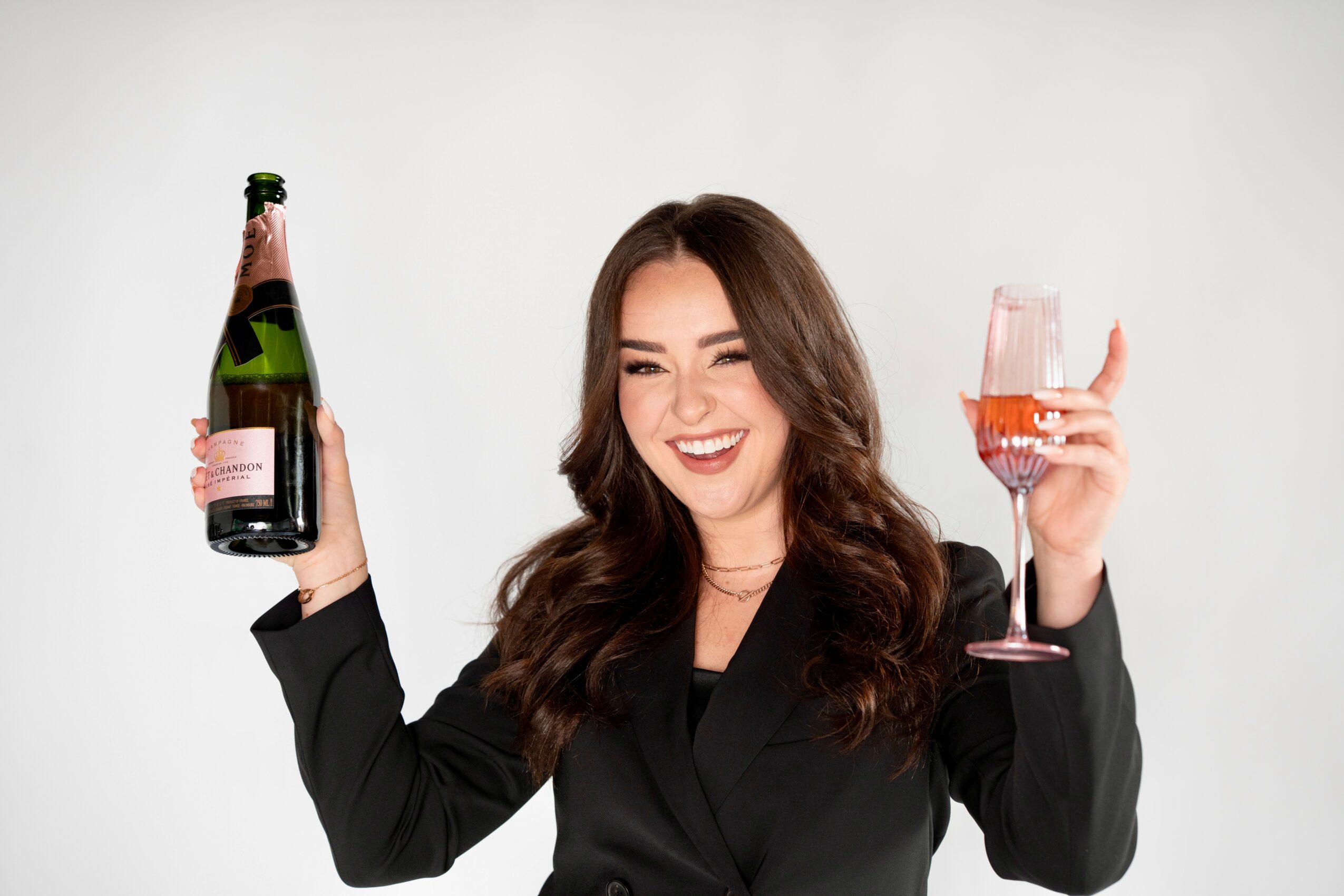 Woman with long brown hair smiling, holding a champagne bottle in one hand and a glass of pink drink in the other, against a white background.