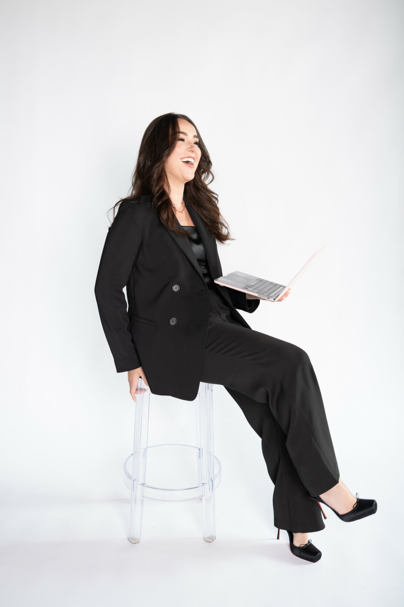 Woman sitting on a transparent chair, holding a laptop, smiling, wearing a black suit, against a plain white background.