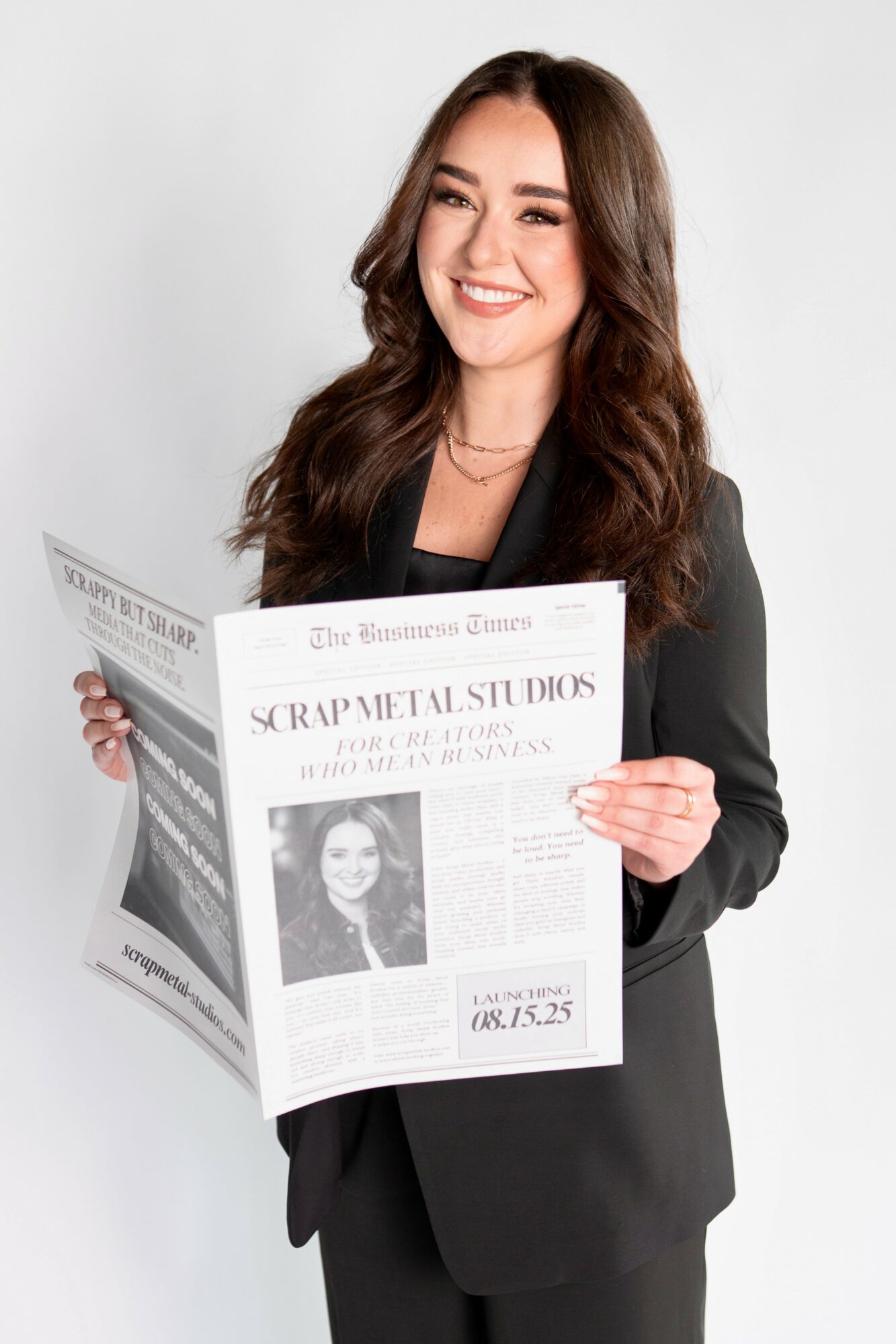 Smiling woman with long brown hair in a black blazer holding a newspaper, standing against a white background.
