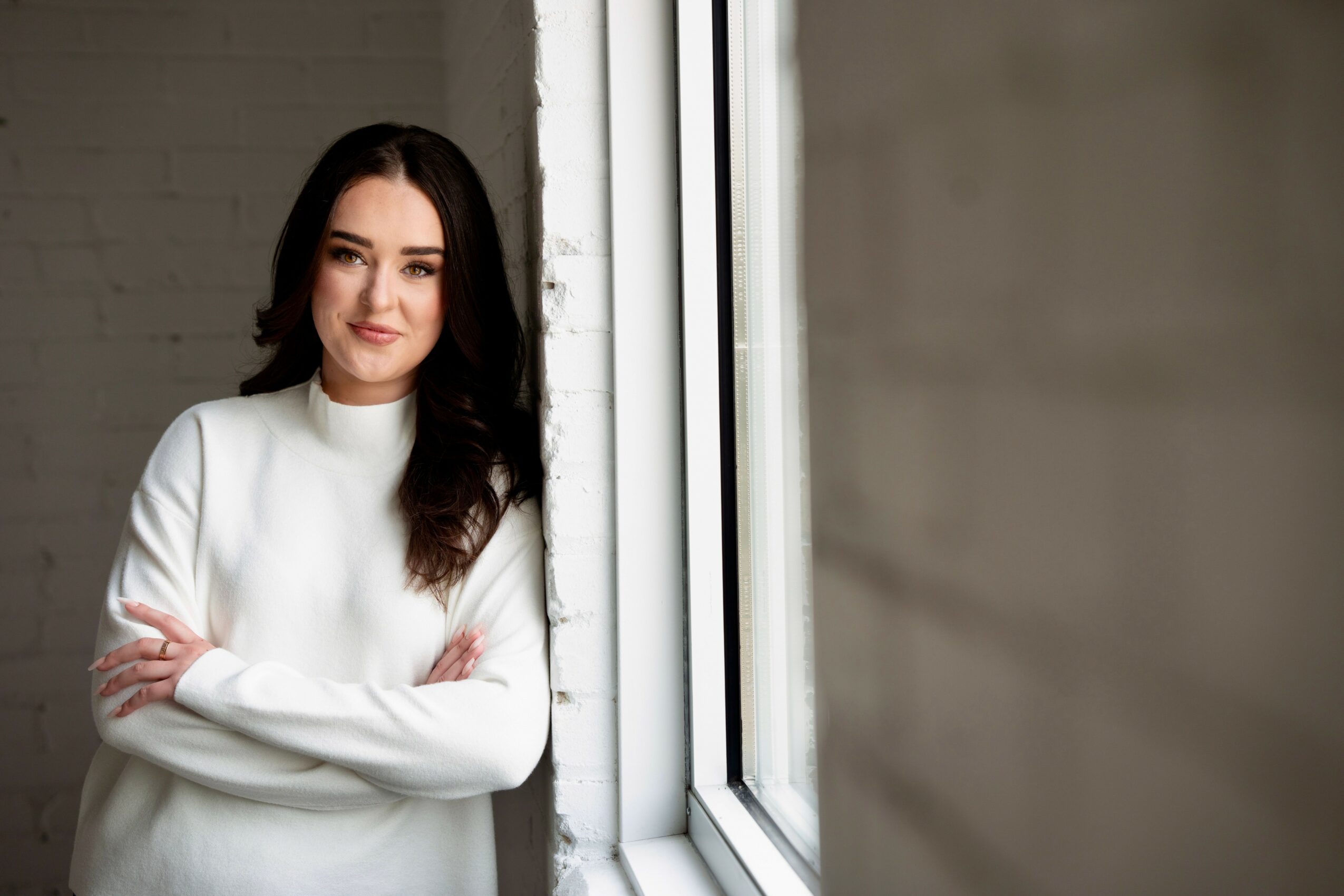 Woman with dark hair and white sweater standing by window, arms crossed, smiling, indoor setting