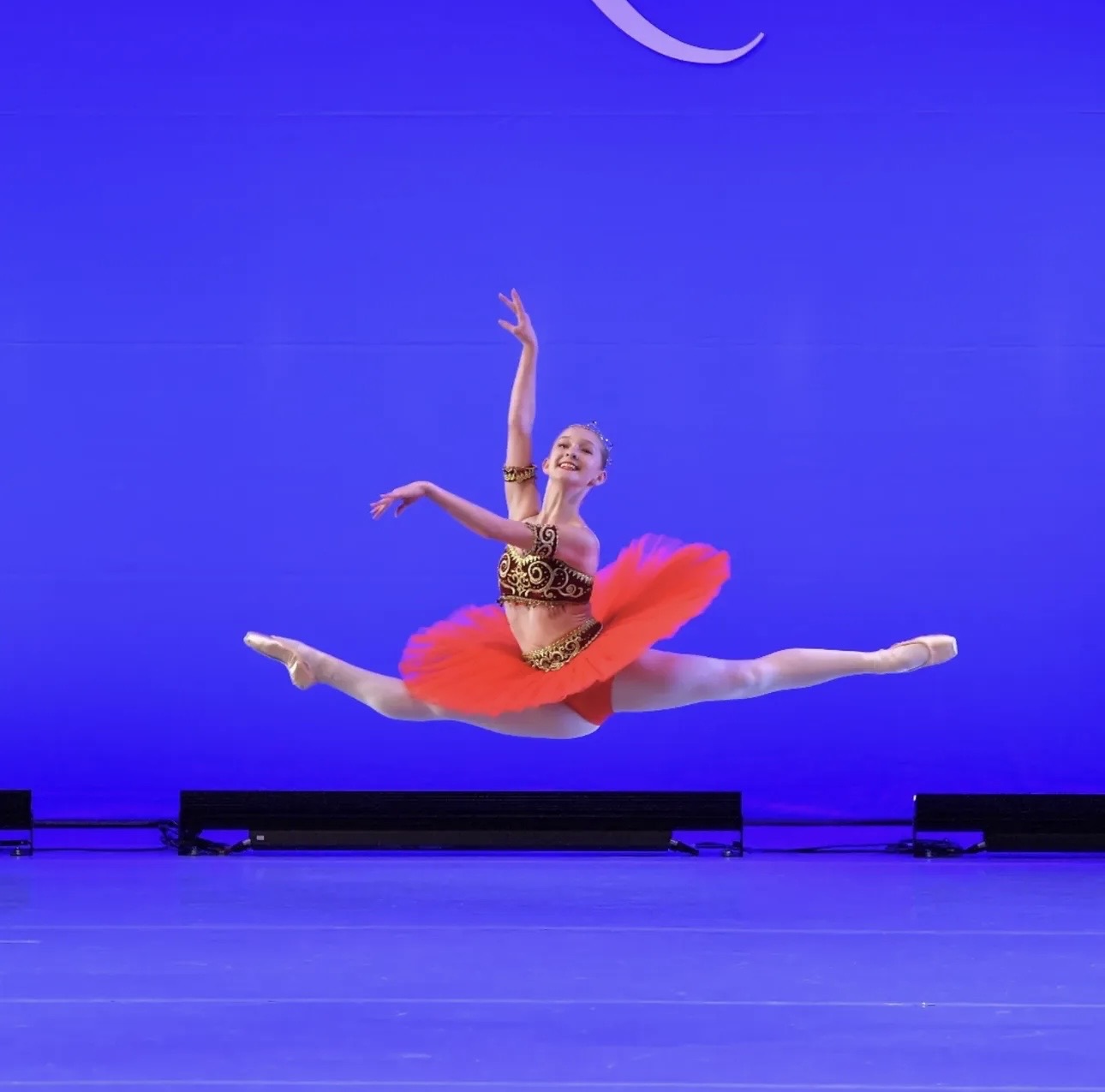 Ballet dancer in a red tutu performing a split leap on stage with a blue background.