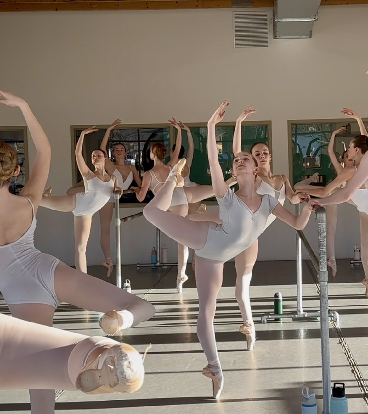 Ballet dancers practicing in a studio, some in poses with arms raised and one on one leg, sunlight streaming in.