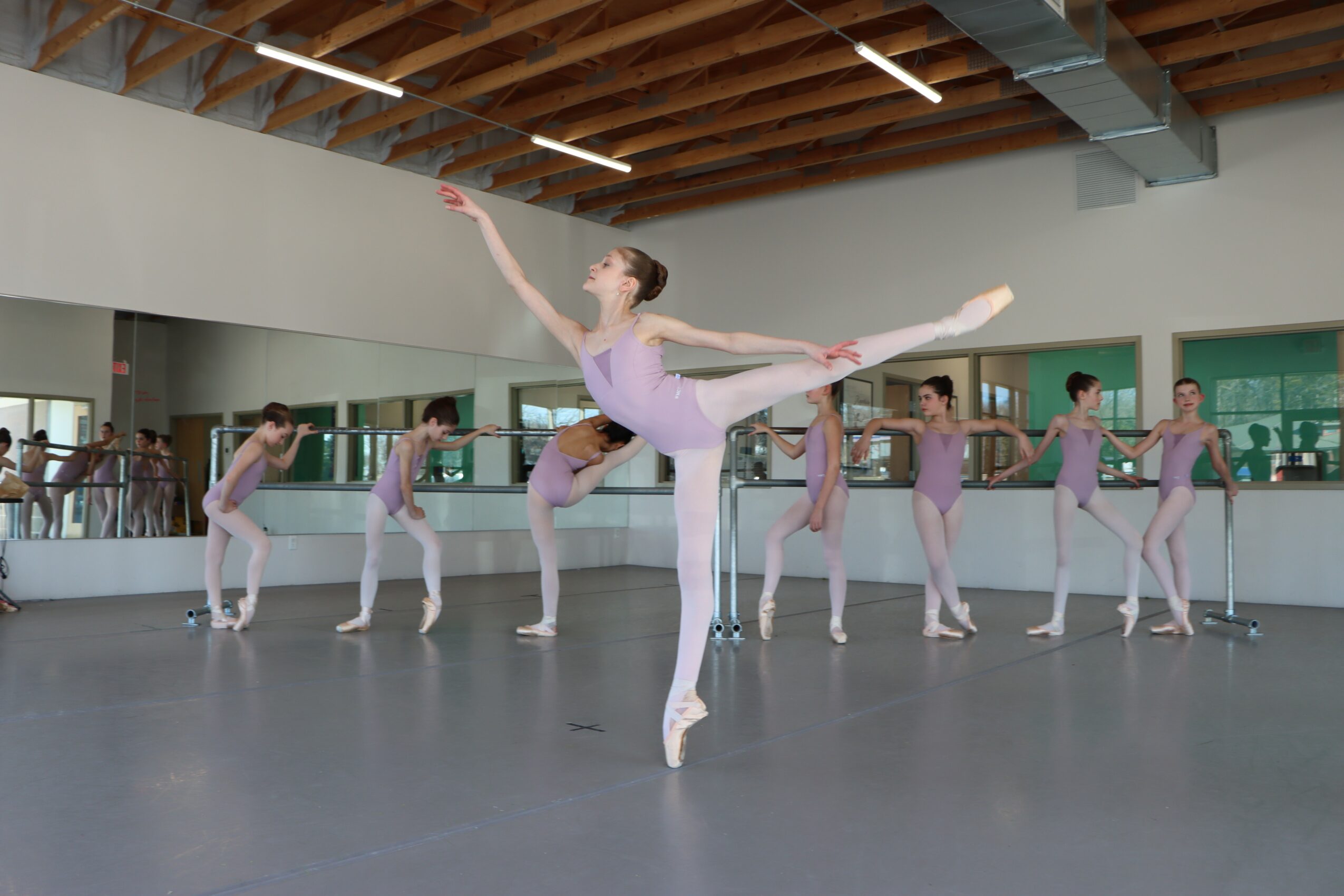 Ballet dancers practicing in a studio with mirrors, wooden ceiling, and large windows, with one dancer in front performing a leap.