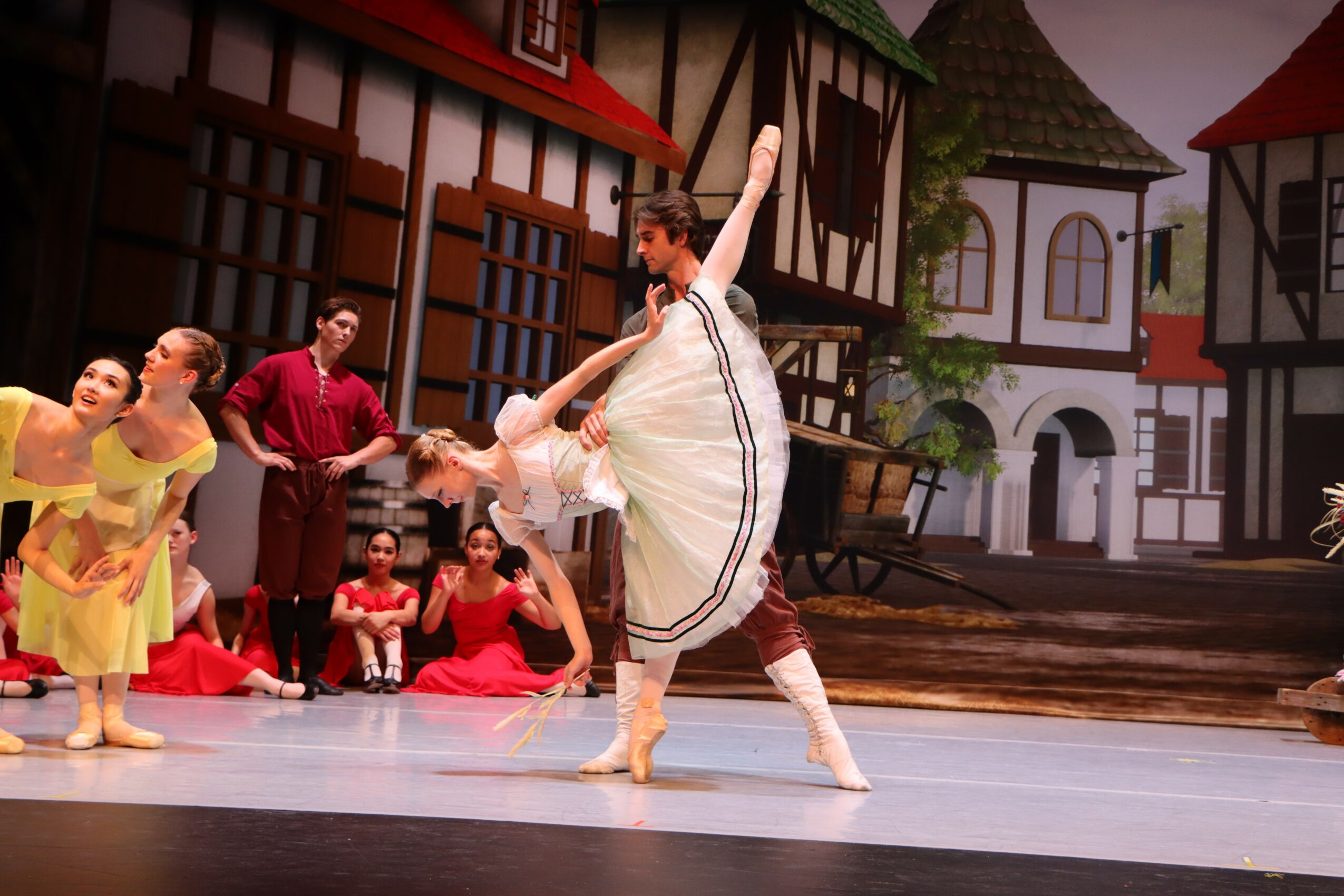 Group of children performing a dance on stage with a village backdrop, some in colorful costumes, one in a white dress, others sitting or standing.