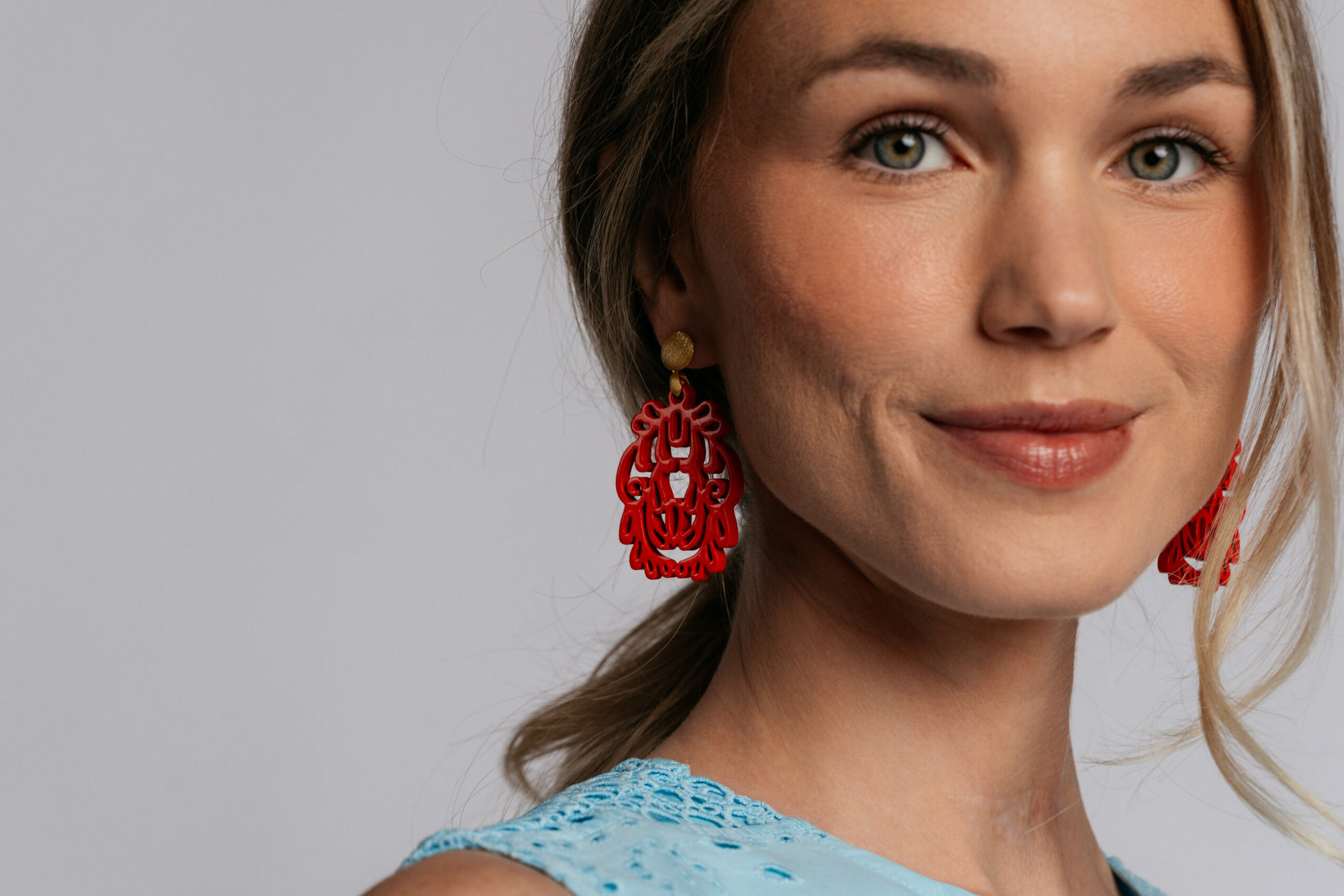 Close-up of a woman with light brown hair, wearing red earrings and a blue top, smiling softly.