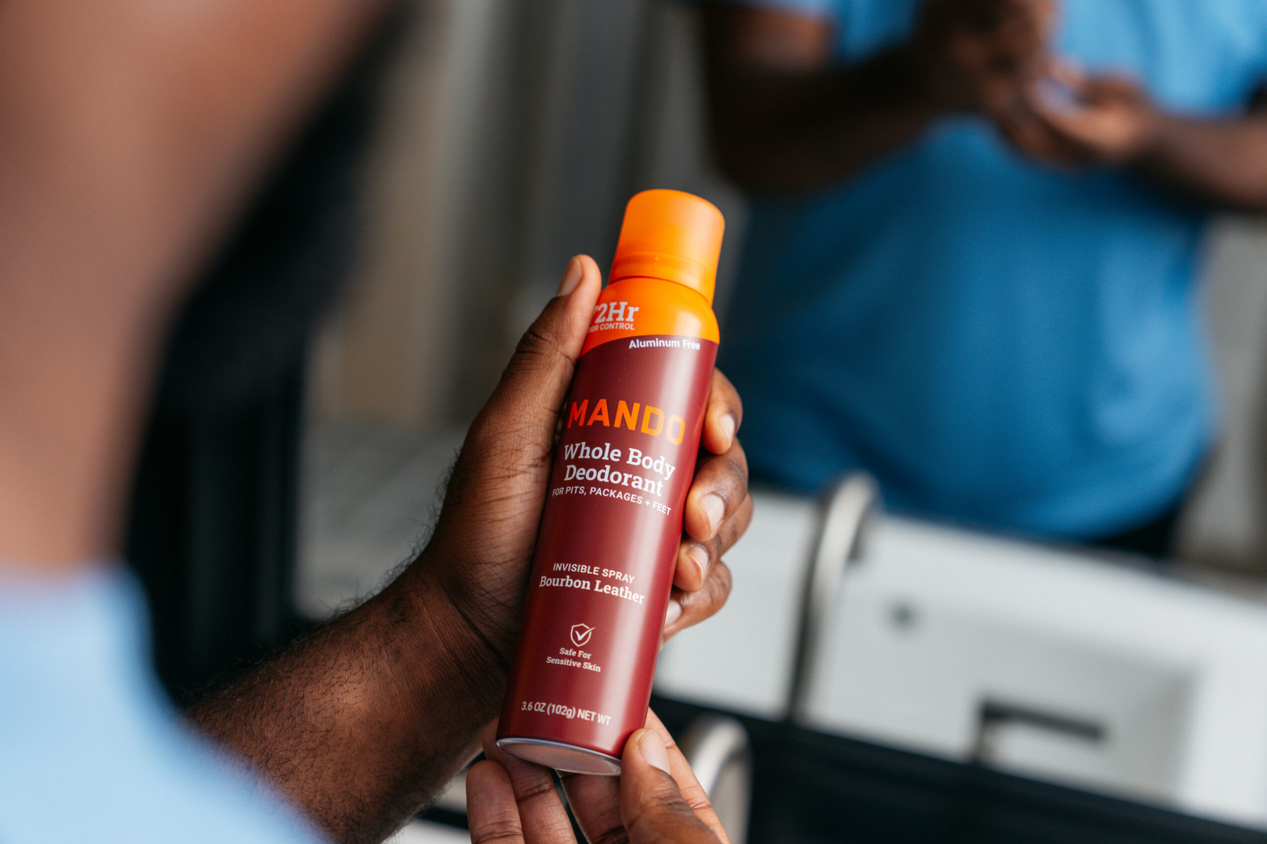 Person holding a red and orange spray bottle labeled 'MANI' in a store, with another person in the background wearing a blue shirt.