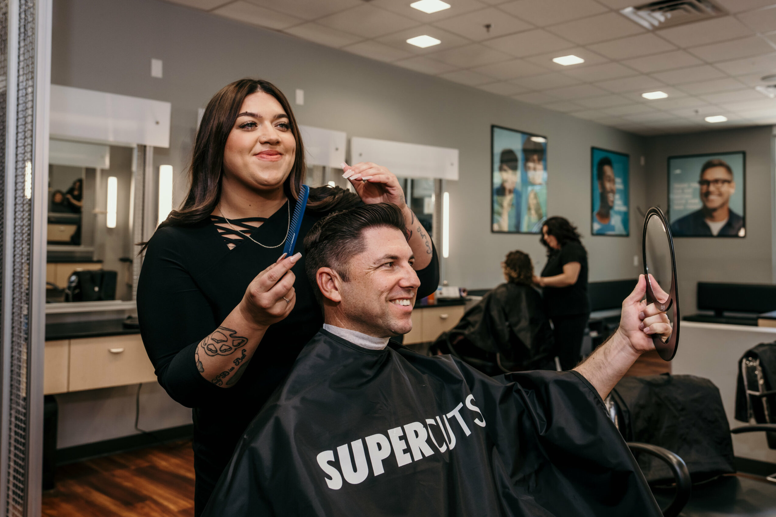 Woman smiling while cutting man's hair in a salon, man looking at mirror, other clients in background.