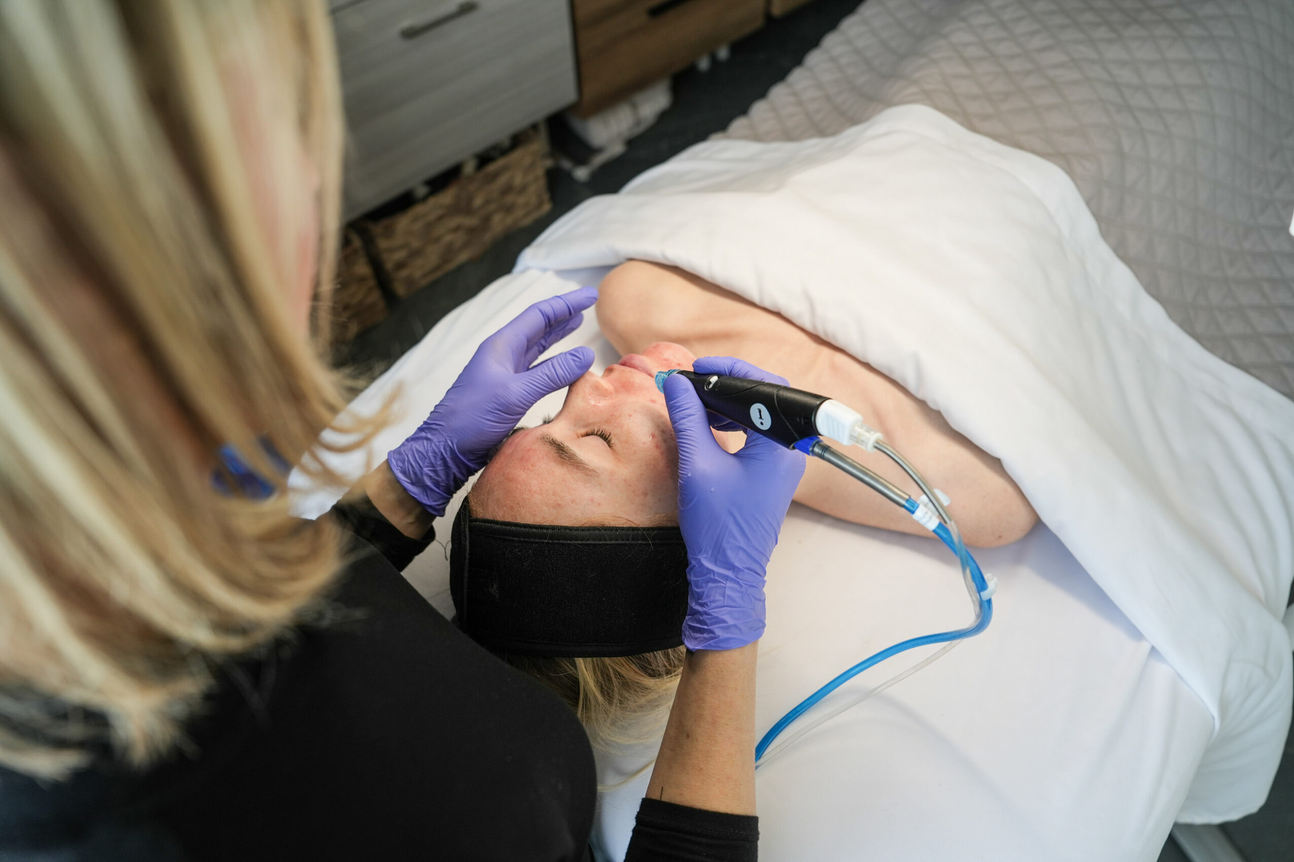 Person receiving a facial treatment with a device on their face, lying on a bed covered with a white sheet.