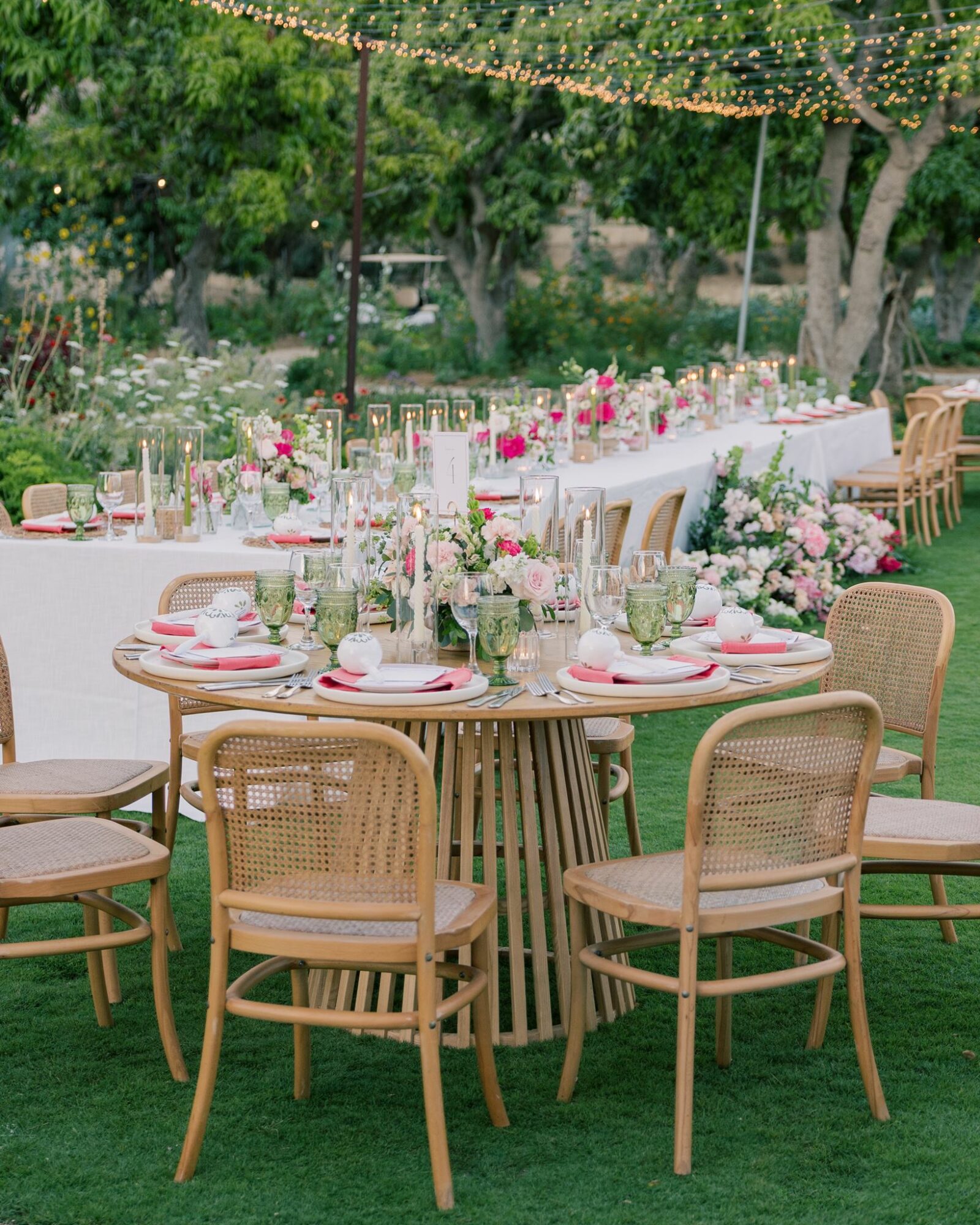 Outdoor event setup with round and long tables decorated with flowers, candles, and tableware, under string lights and trees.