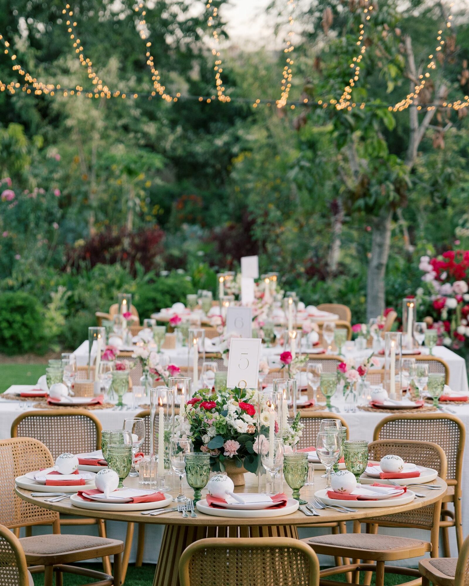 Outdoor garden table set for a celebration with floral centerpieces, candles, and place settings, surrounded by greenery and string lights.