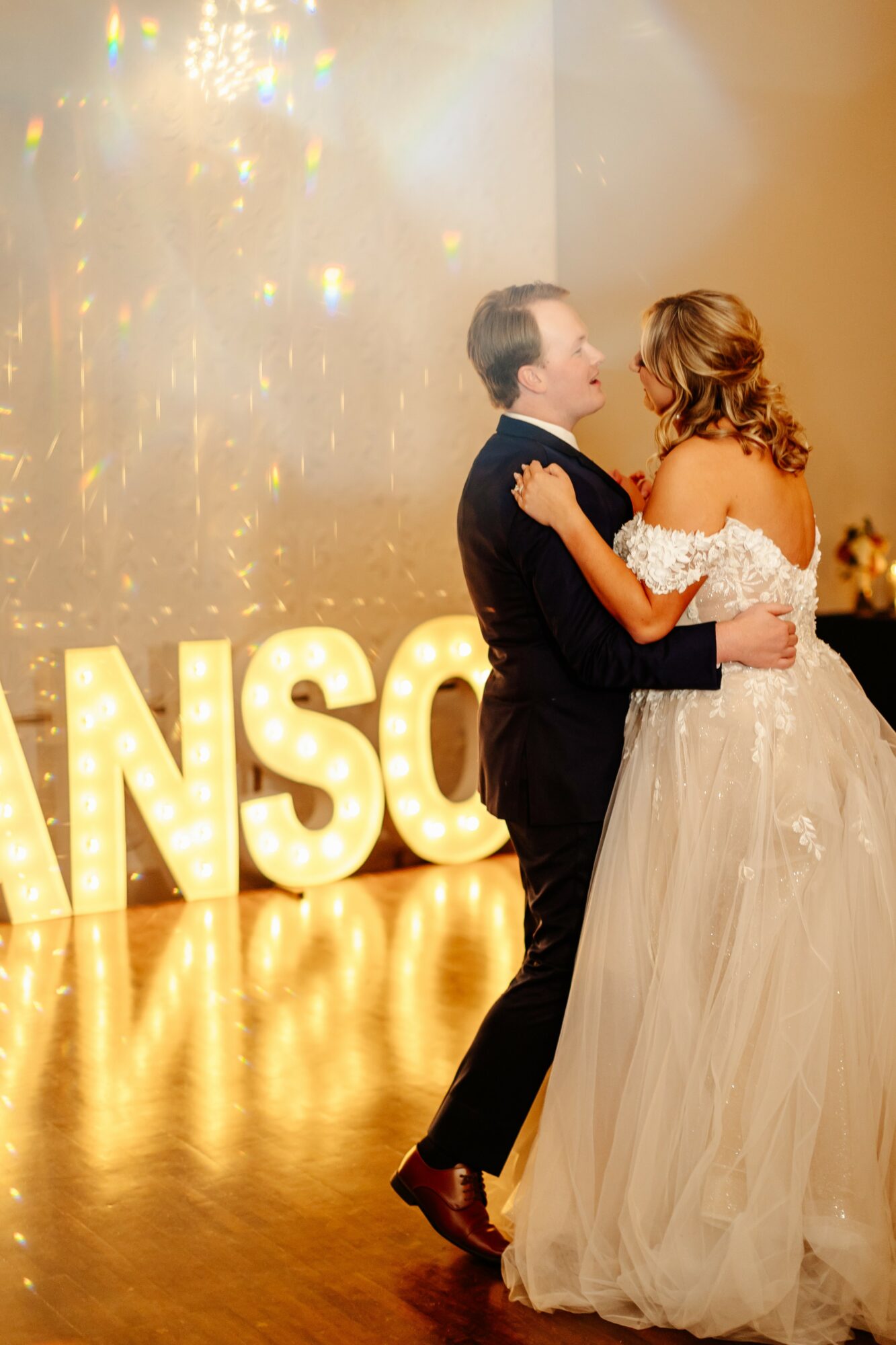 A couple dancing at a wedding reception, with large illuminated letters spelling 'MANSO' in the background.