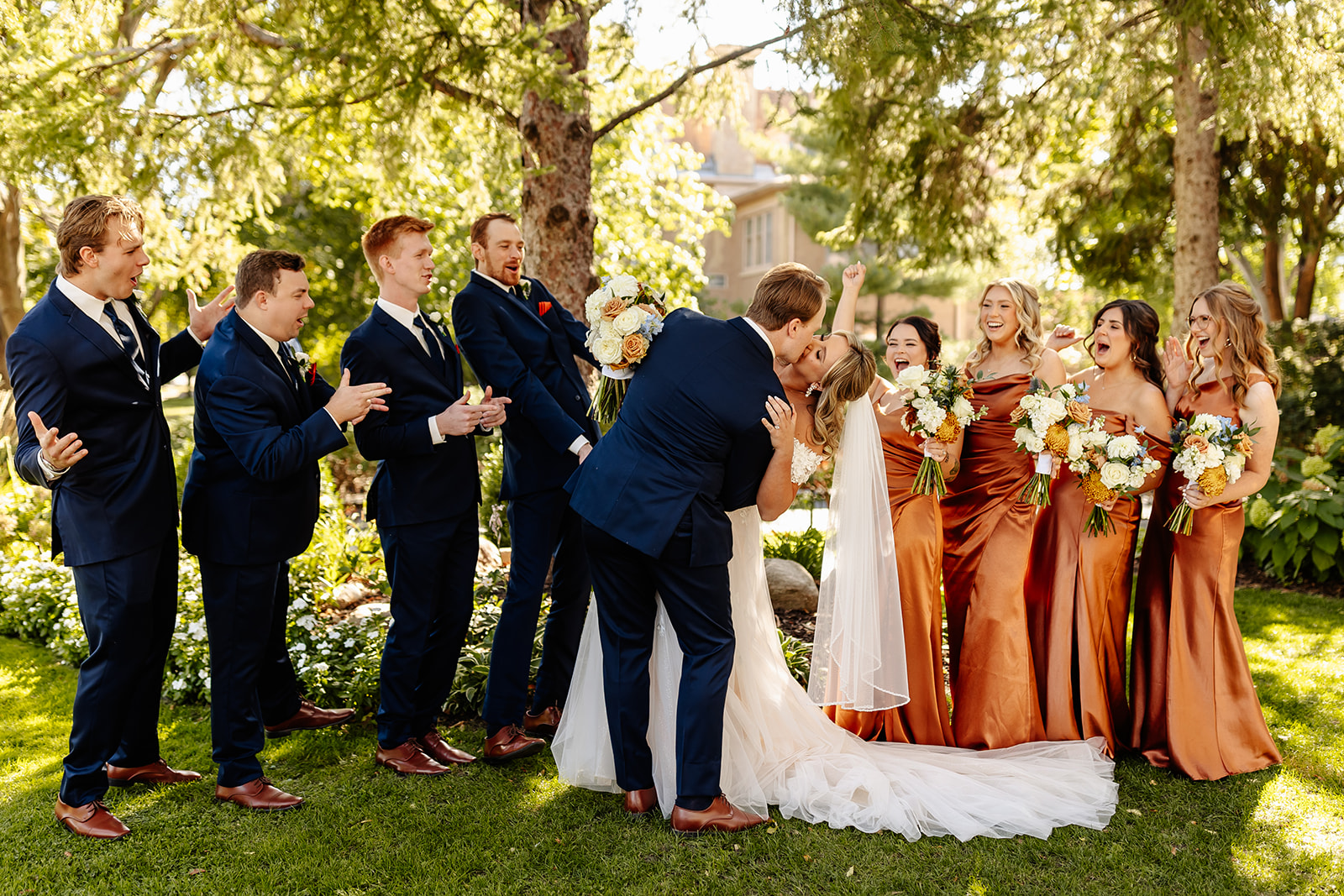 Groom kisses bride while bridesmaids and groomsmen stand outdoors among trees, sunlight, and greenery, celebrating wedding.