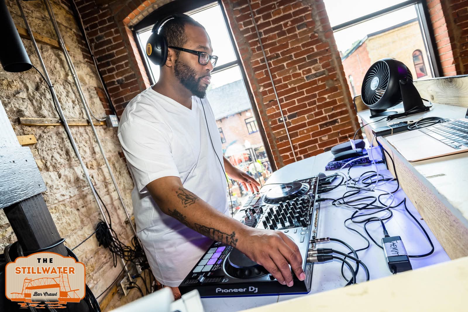 DJ wearing headphones operates equipment in a brick-walled room with large windows, sunlight, and a laptop on the table.