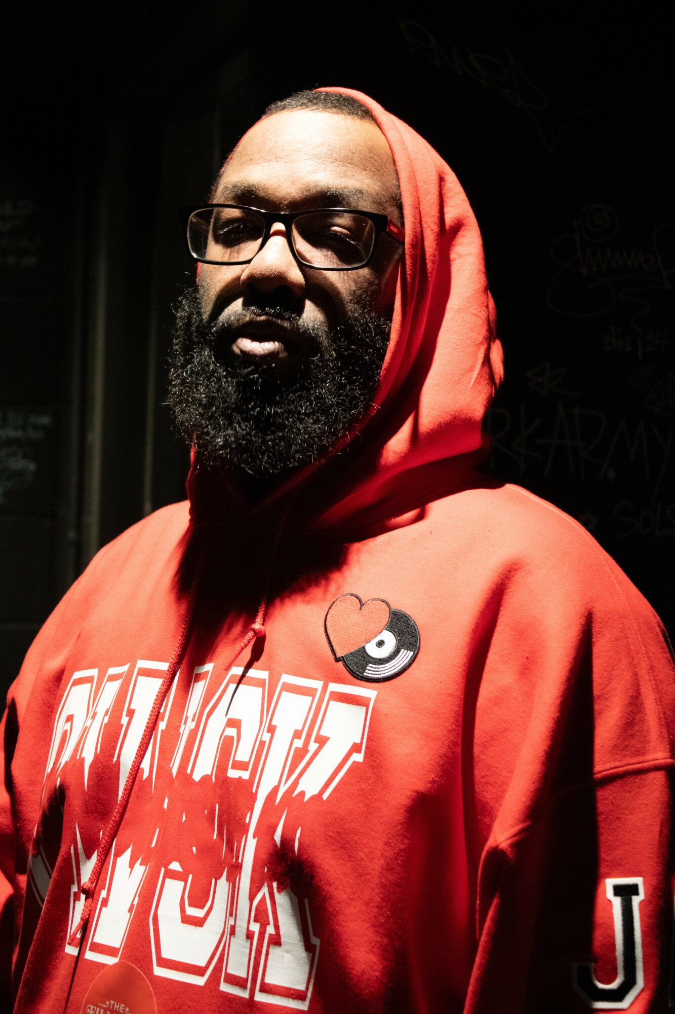 Man with glasses and beard wearing a red hoodie, facing slightly to the left, dark background.