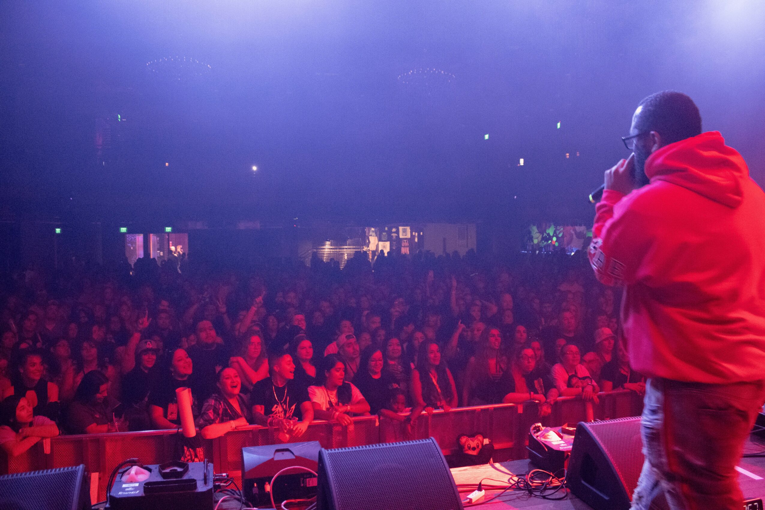 Performer on stage facing a large audience in a dark venue with purple lighting, wearing a red hoodie and glasses.