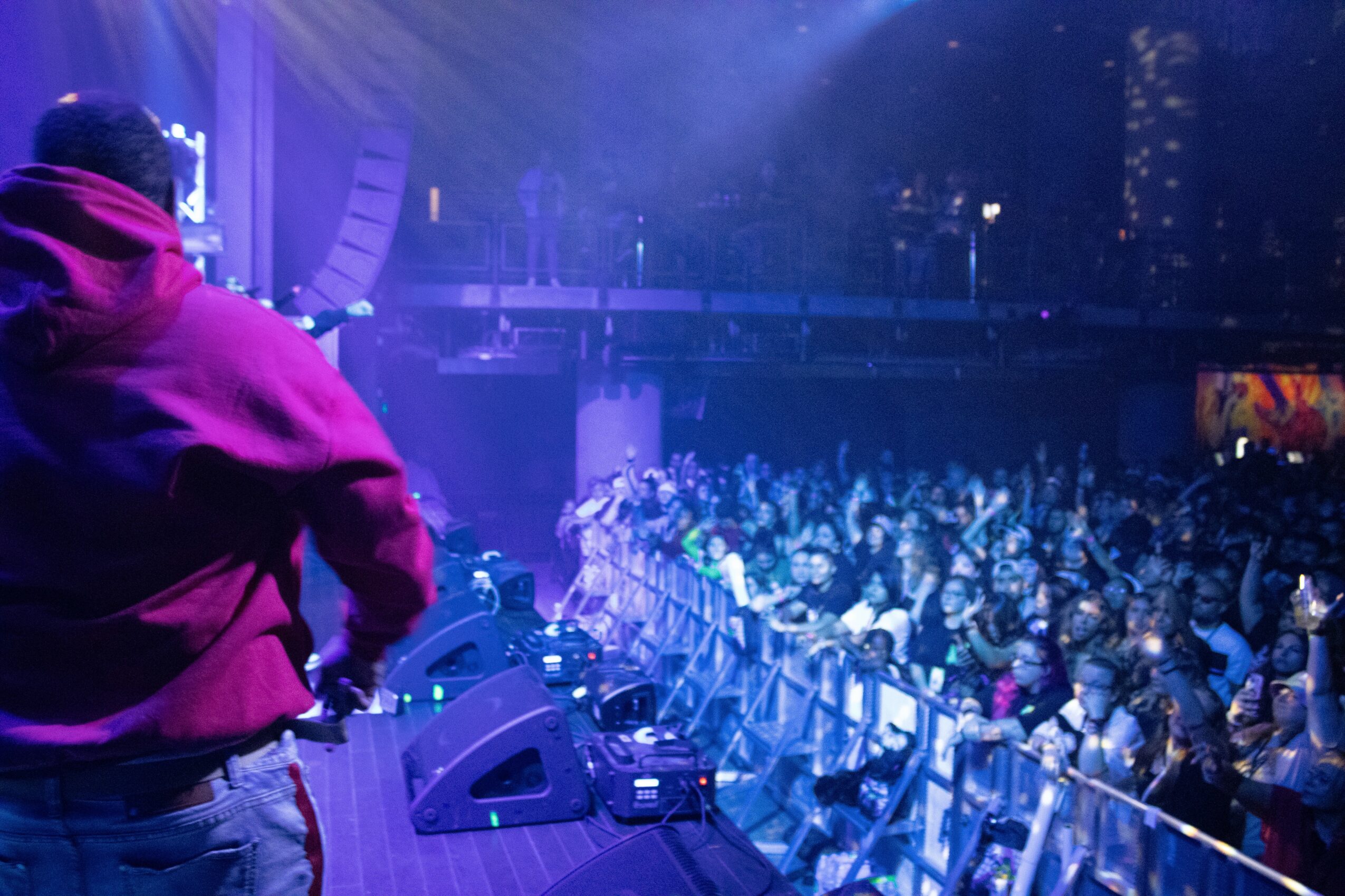 Person in a red hoodie facing a large crowd at a concert or event, stage lights illuminate the scene.