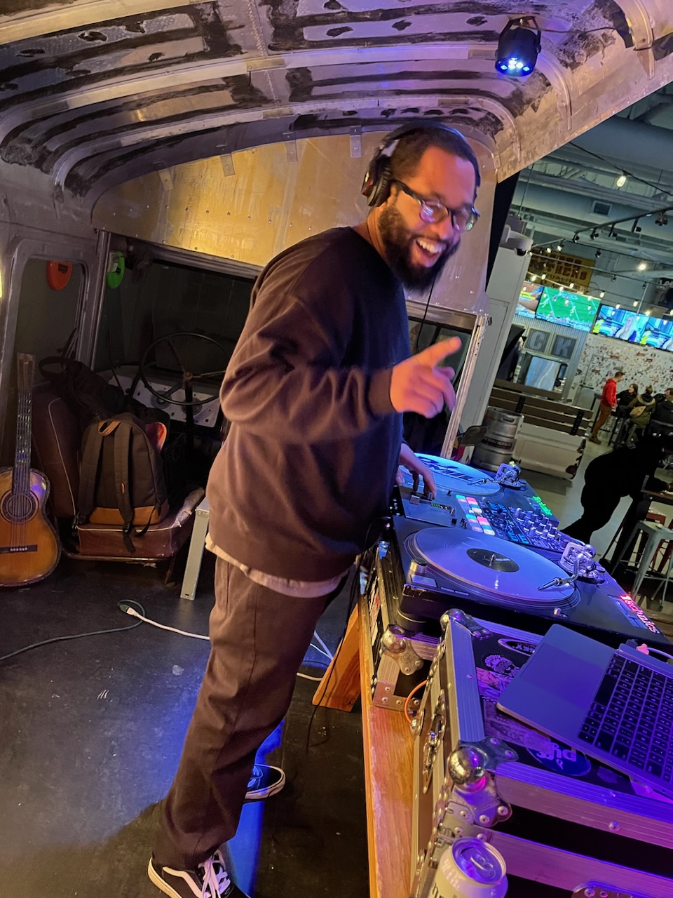 Person with glasses and headphones smiling and pointing at DJ equipment in a bar or club setting.