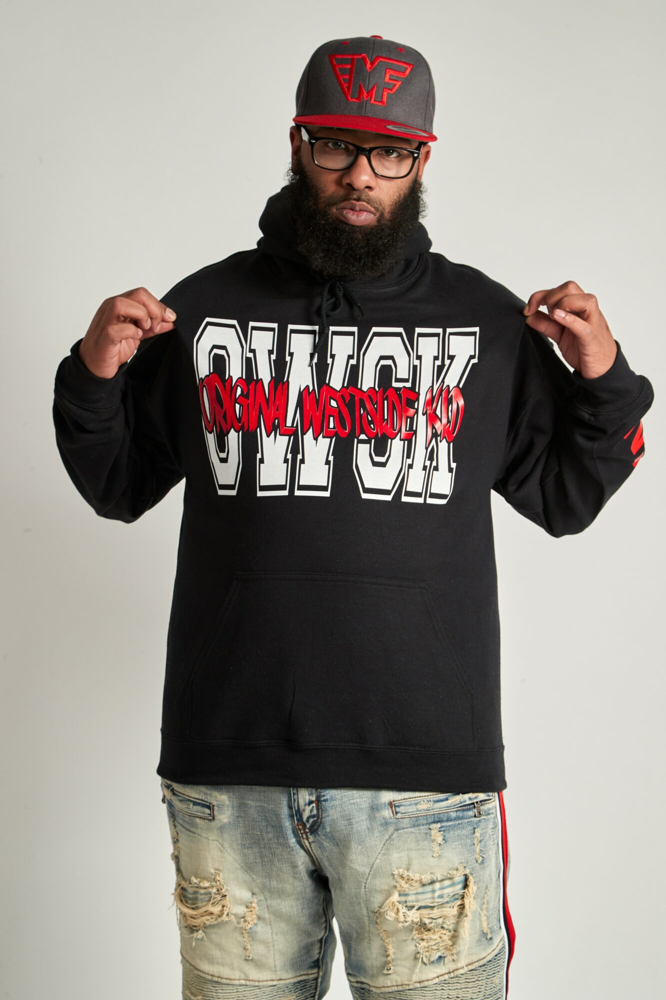 Man with glasses and a beard wearing a black hoodie, red cap, and ripped jeans, standing against a plain background.