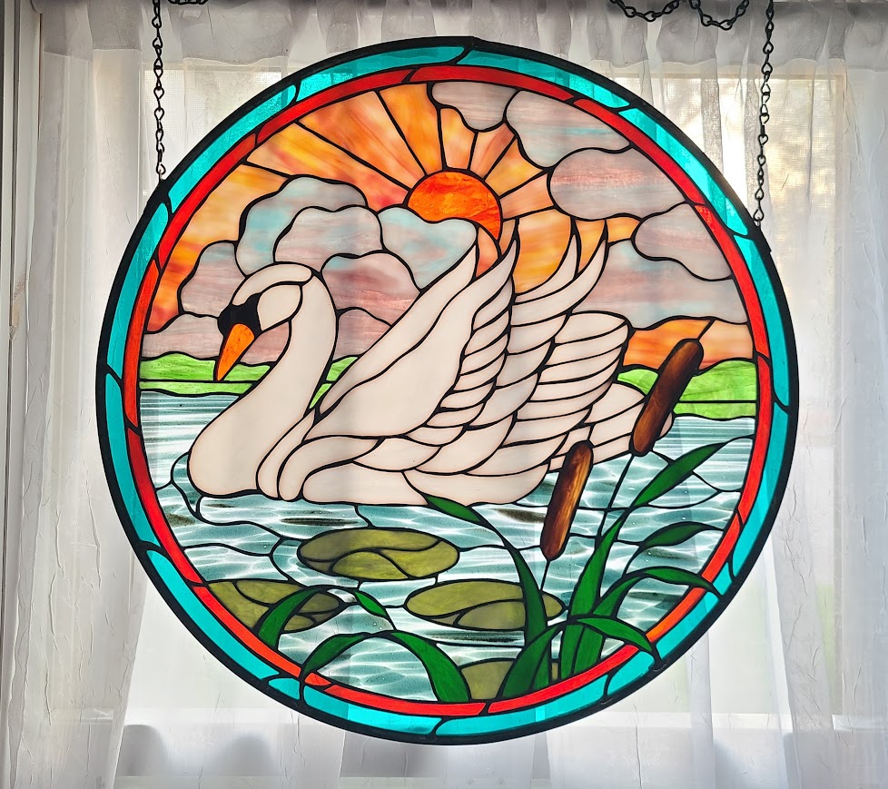 Stained glass window depicts two swans on water with a sunset, clouds, and reeds in the background.