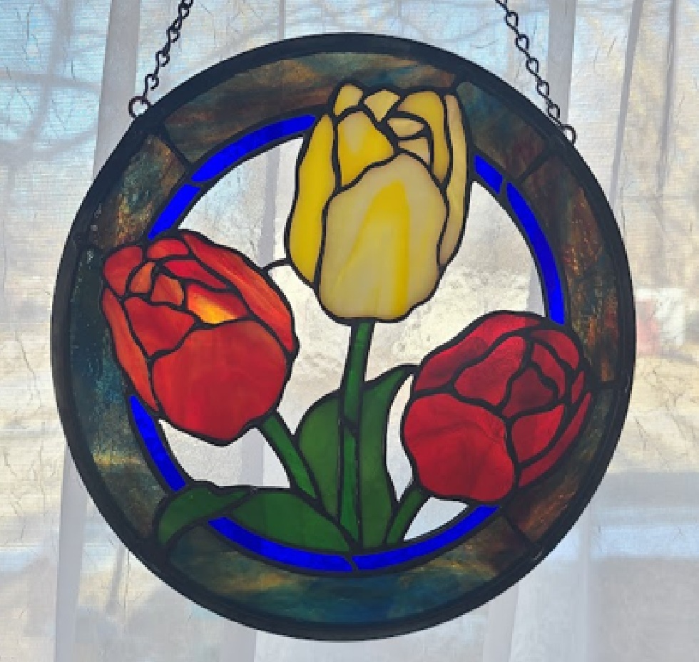 Stained glass circular window with three tulips, two red and one yellow, against a light background.