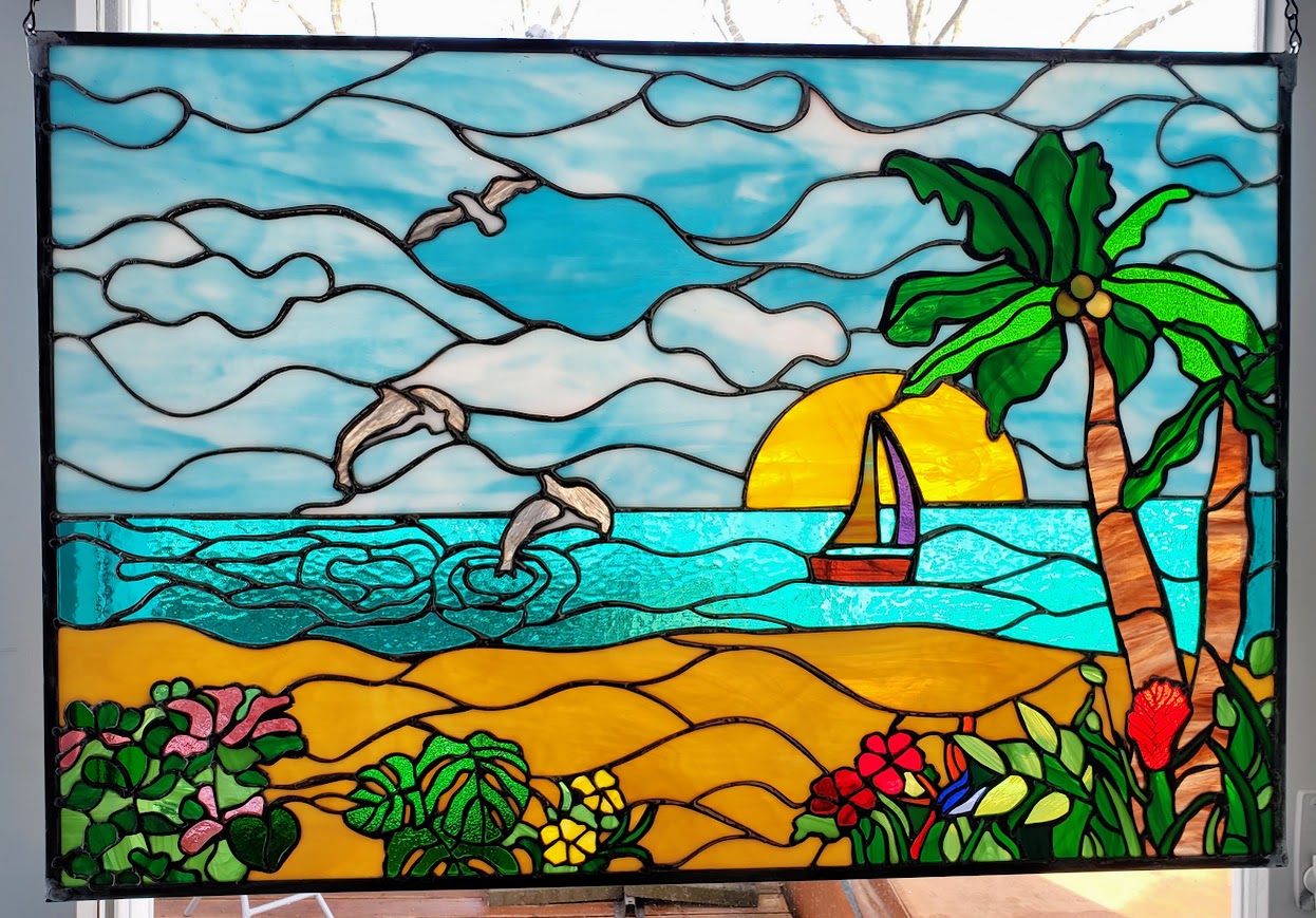 Colorful stained glass scene of a beach with palm trees, seagulls, sailboat, sun, and ocean waves, with flowers in foreground.