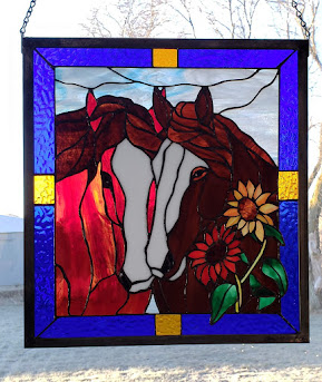 Stained glass window depicting two horses' heads with flowers, framed in blue and yellow, outdoors with snow and trees.