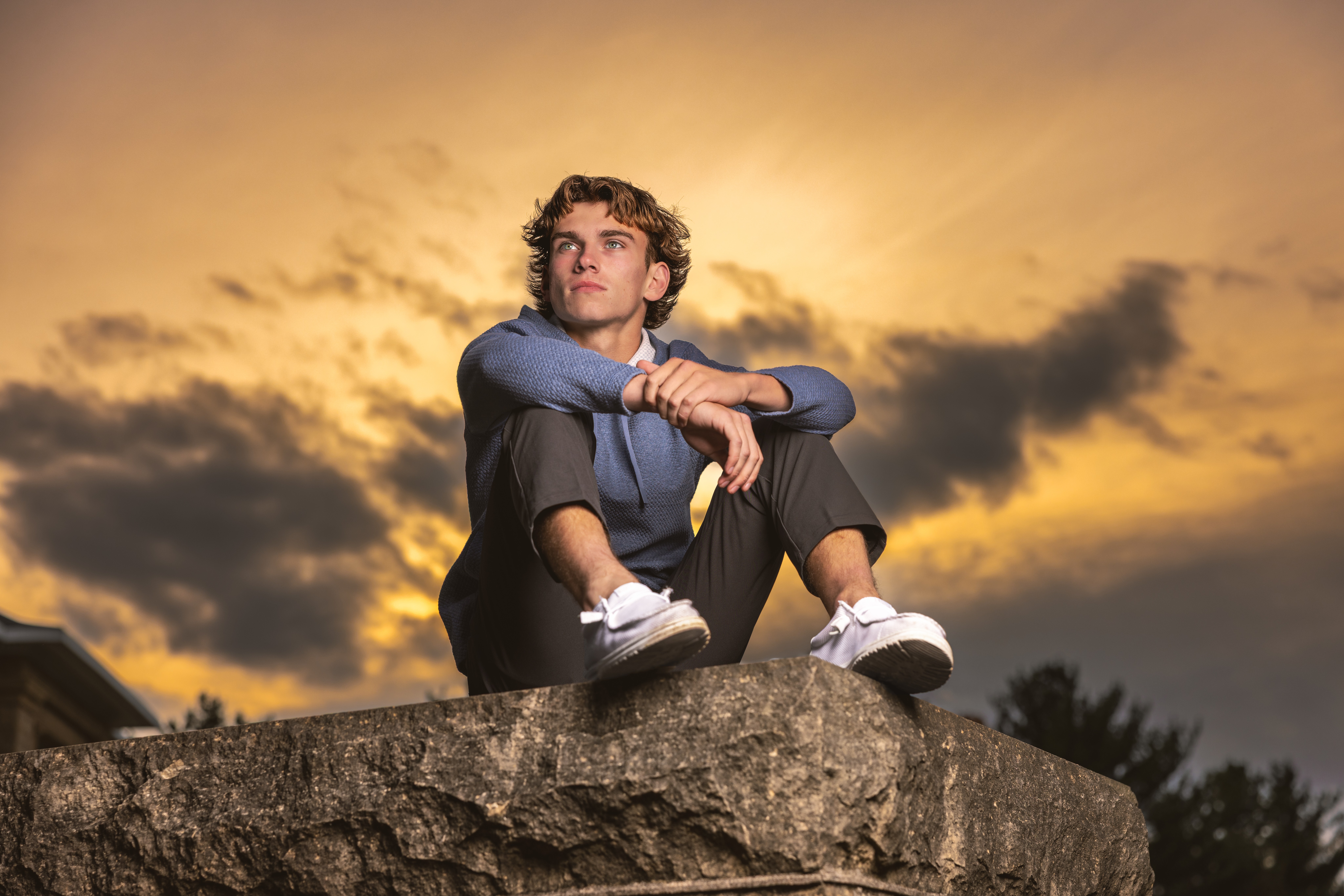 Young man sitting on a stone ledge with a sunset sky in the background, looking thoughtful, arms resting on knees, wearing casual clothes.