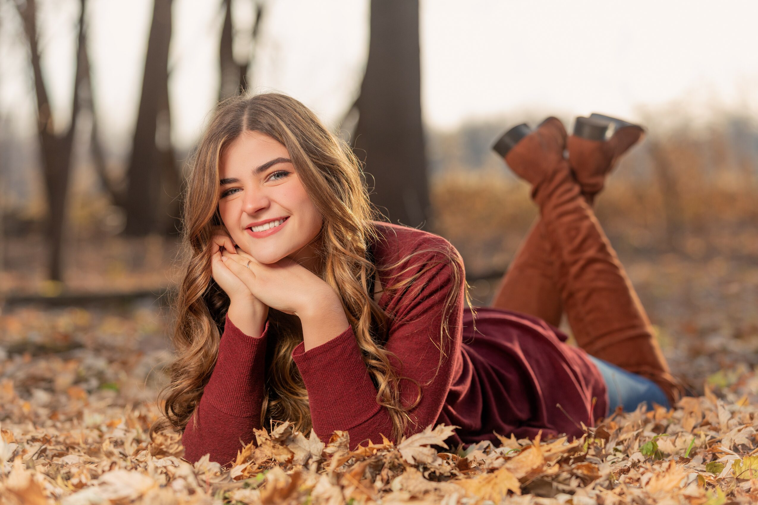 Young woman lying on autumn leaves in a forest, smiling, with her chin resting on her hands, wearing a maroon dress.