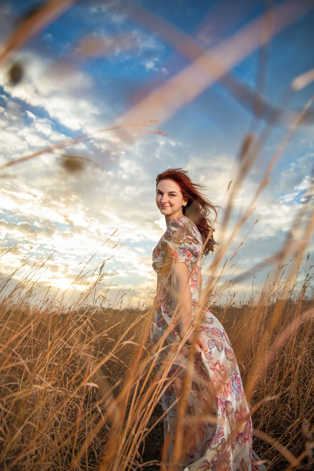 Woman standing in a field of tall grass under a partly cloudy sky, smiling and looking to the side.