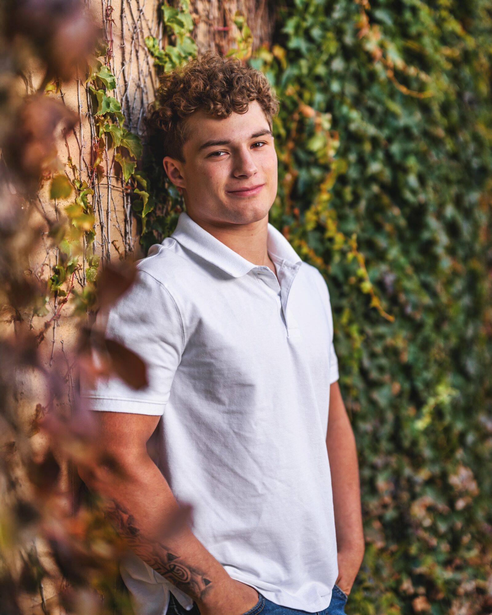 Young man with curly hair leaning against a tree, smiling, surrounded by green and yellow foliage.