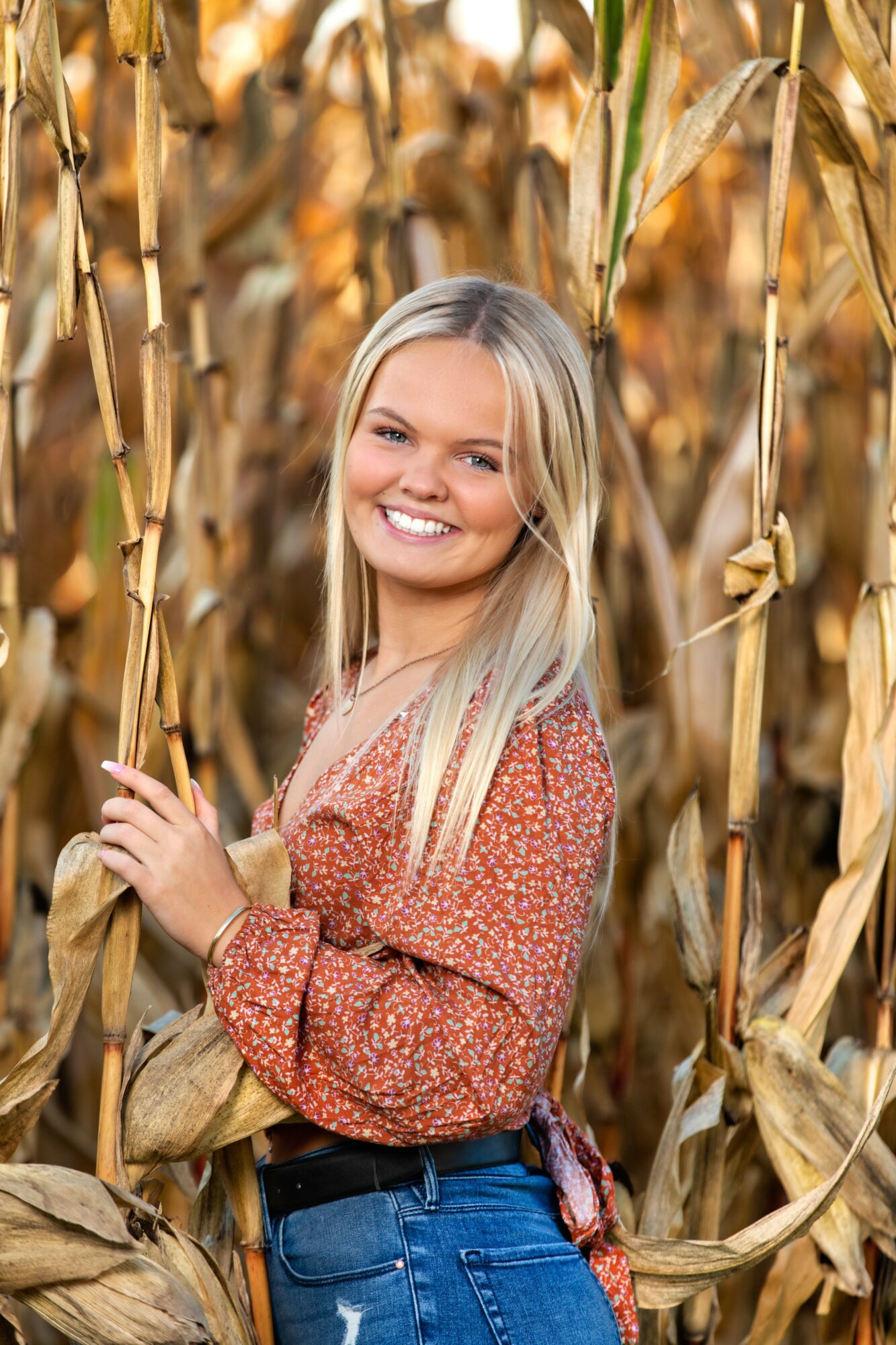 Smiling woman with long blonde hair standing among tall, dry corn stalks in a field.
