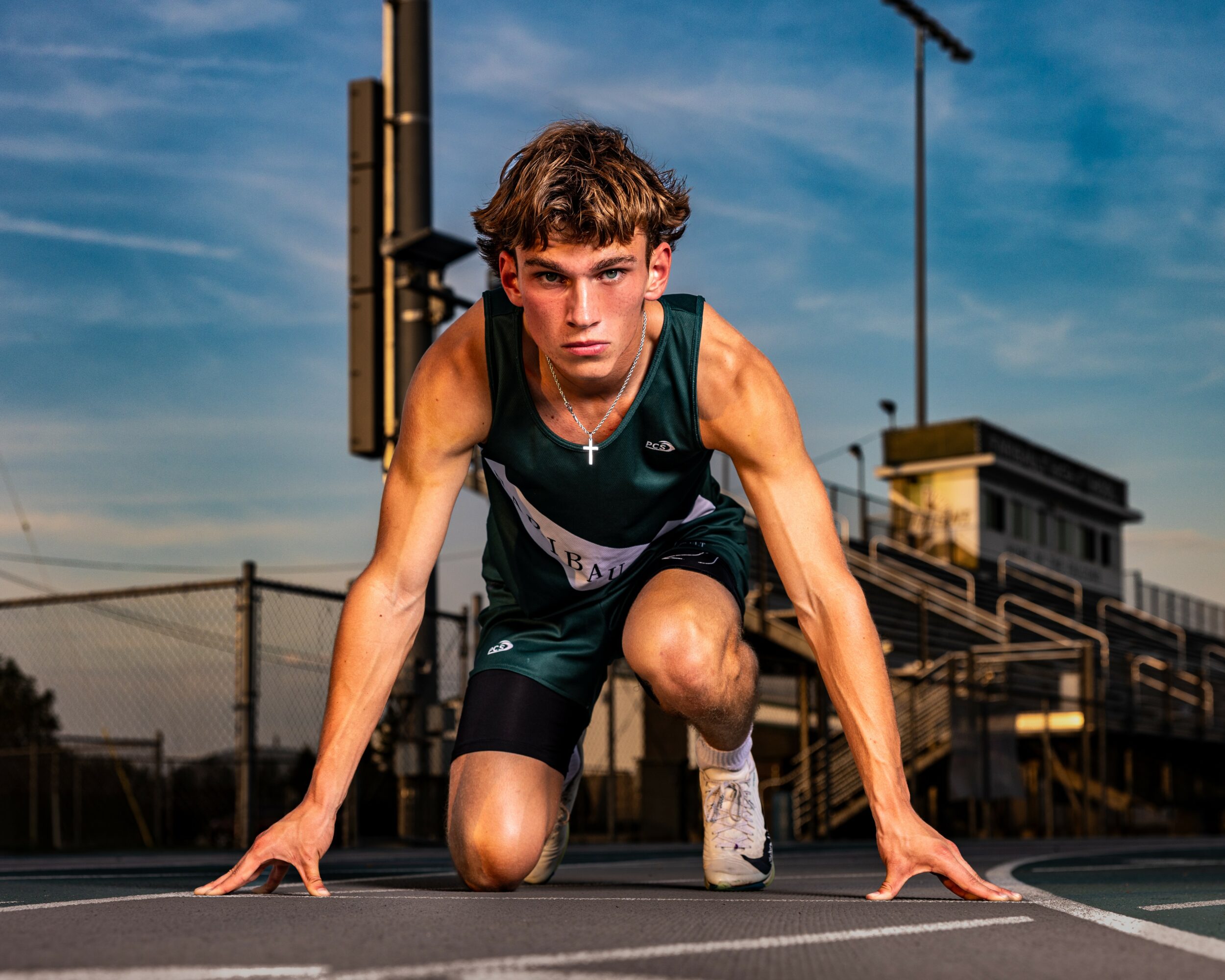 Young male athlete in starting position on a track, outdoors, with stadium and sky in background.